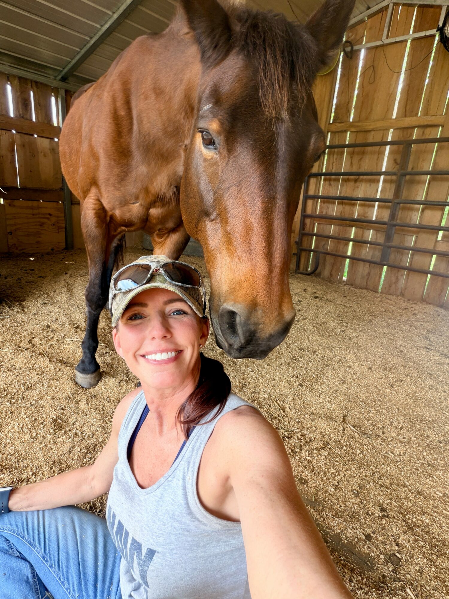 Woman smiling in a barn with a horse behind her, wooden walls and dirt floor visible.