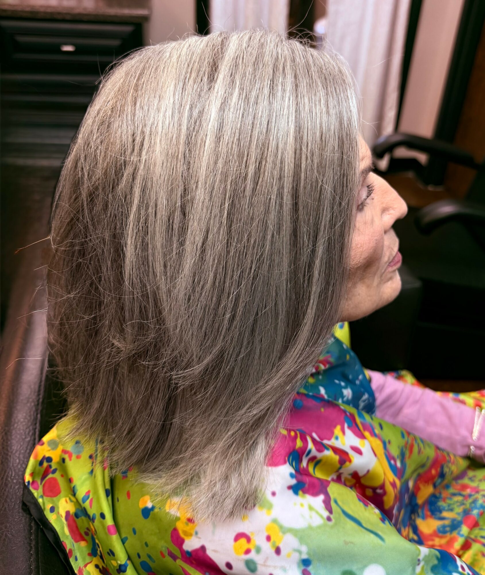 Side view of a woman with shoulder-length gray hair, sitting in a chair wearing a colorful garment.