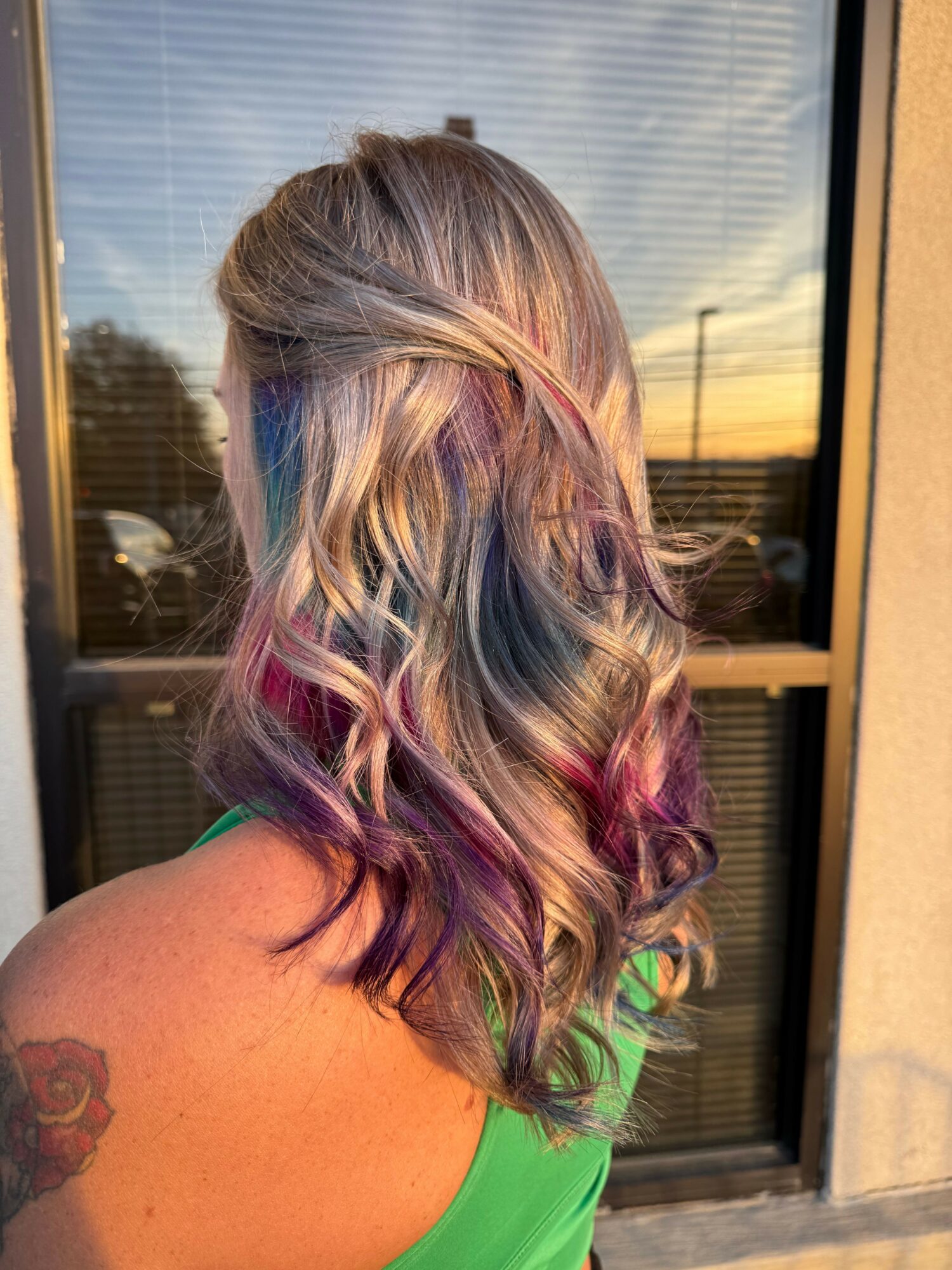 Person with multicolored wavy hair, wearing a green top, standing outside near a window, with a tattoo on their arm.