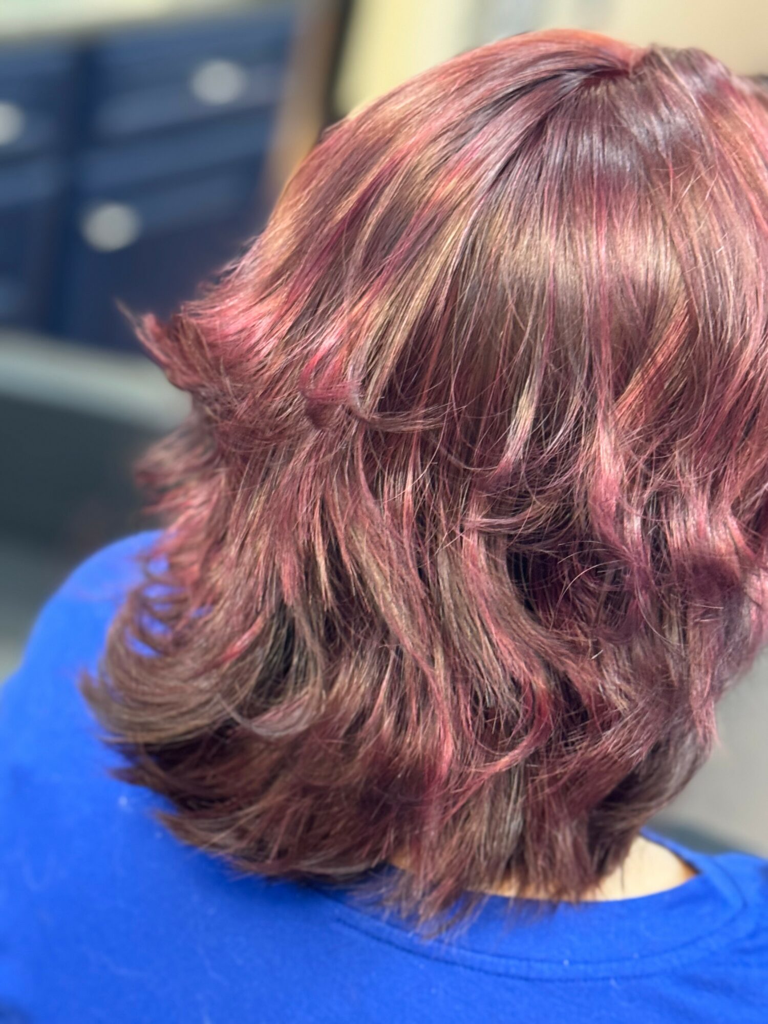 Back of a person's head with shoulder-length, wavy, reddish-brown hair, wearing a blue top.