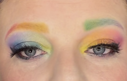 Close-up of eyes with colorful rainbow eyeshadow and eyebrows, makeup blending from yellow to green and blue, with long eyelashes.
