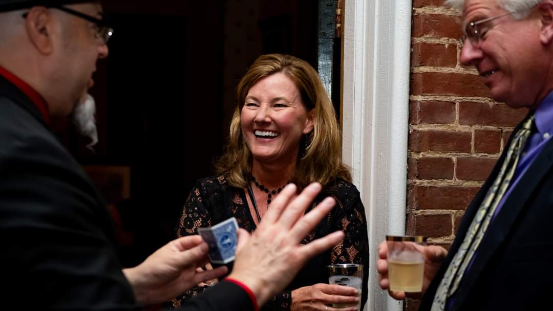 Three people smiling and talking at a social gathering, holding drinks, with a brick wall in the background.
