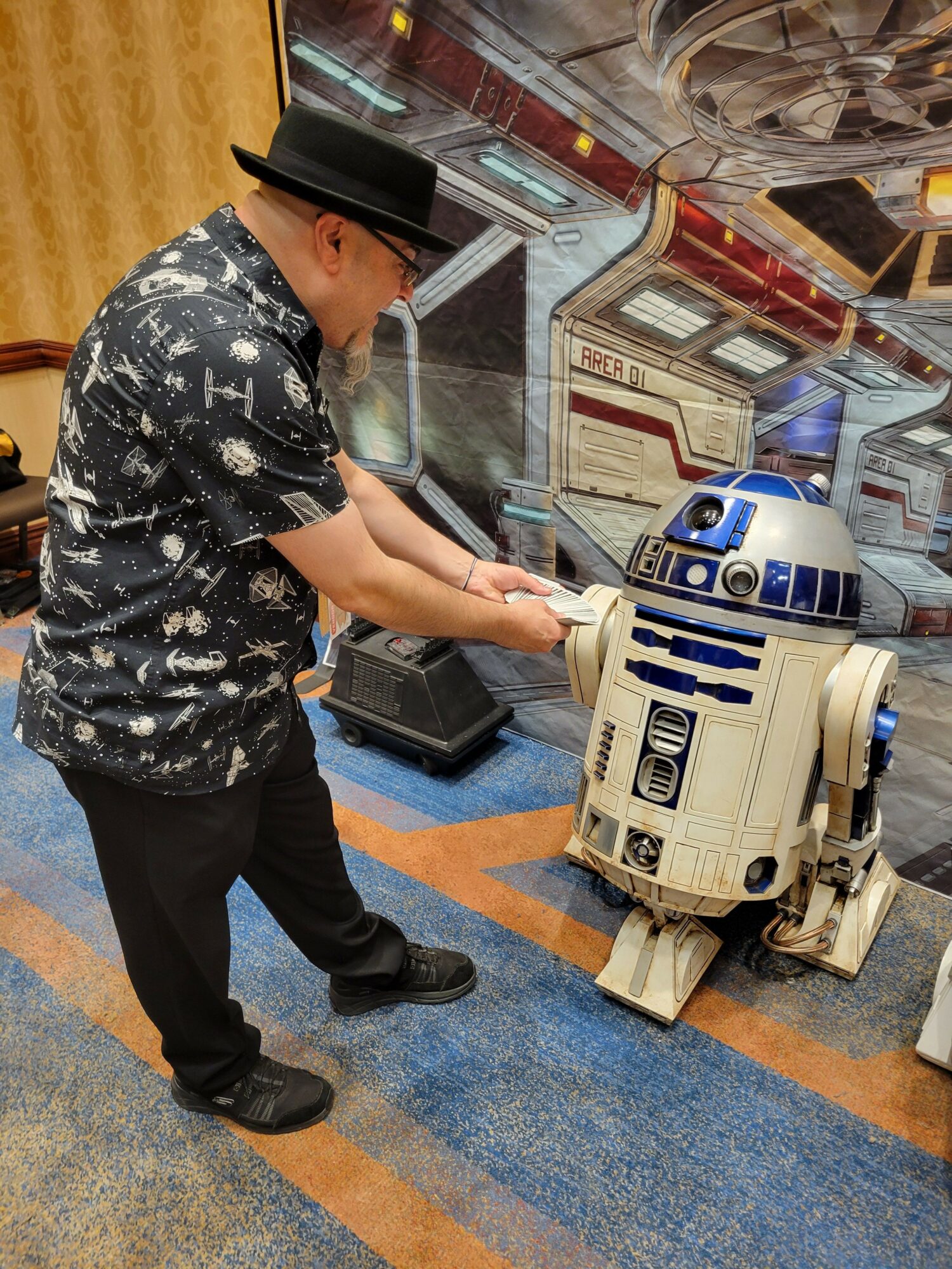 Person dressed as a robot with a black hat, black pants, and a patterned shirt, standing indoors near a display case.