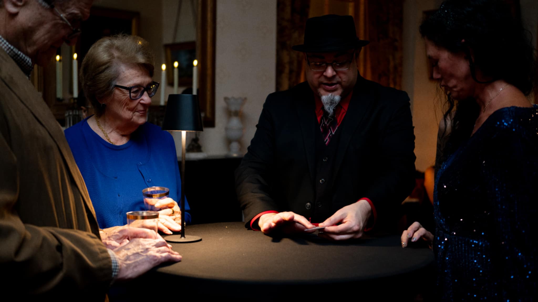 Four people gathered around a table, engaging in a card trick or magic performance, in a dimly lit room.