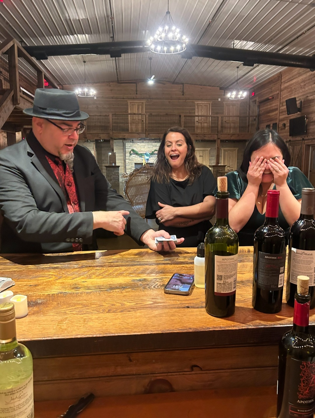 Three people sitting at a wooden bar, two women laughing, one man in a hat and suit, bottles on the bar, rustic interior.