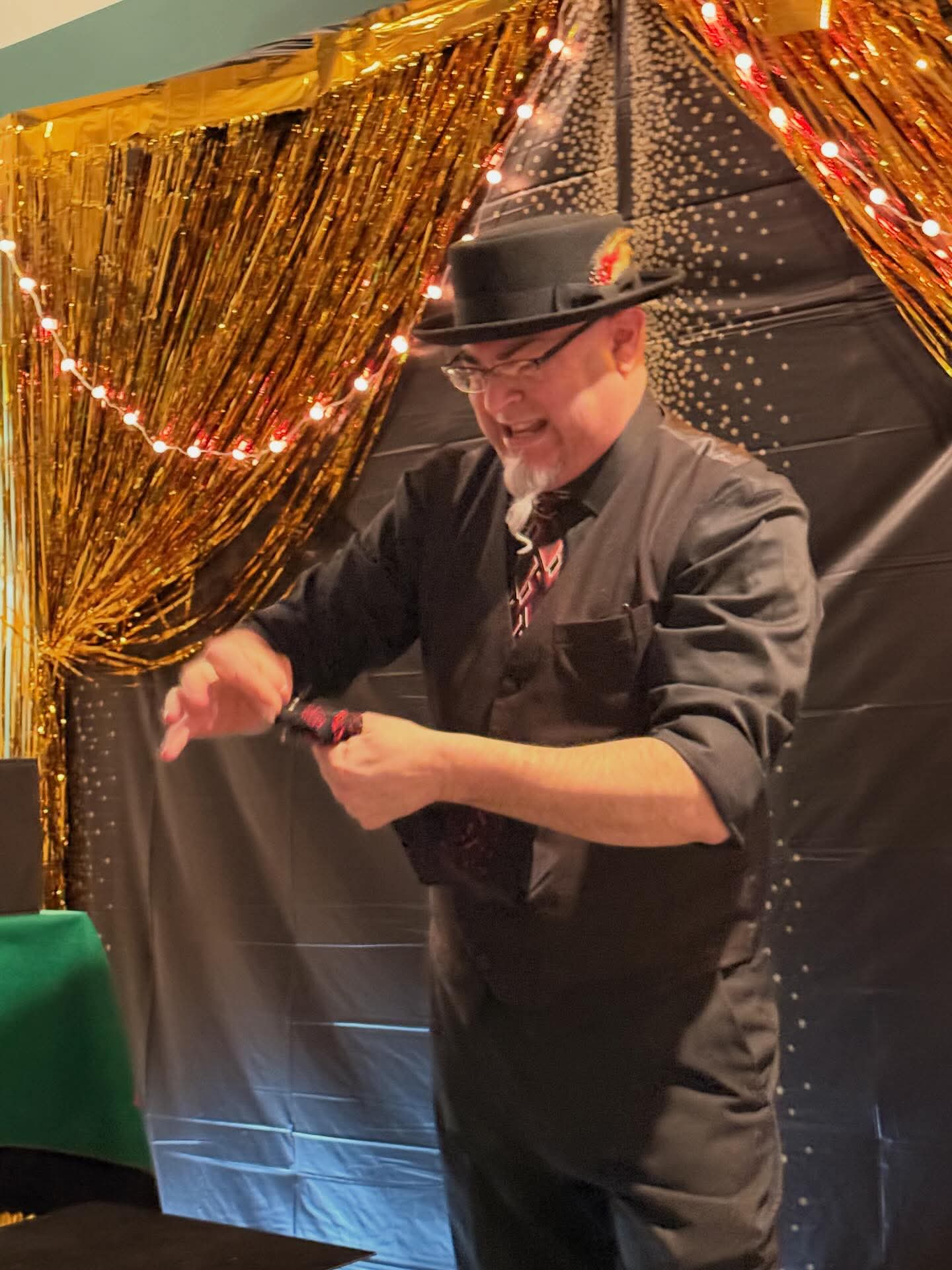 Man in a hat and glasses smiling, holding a card, standing in front of a gold curtain with string lights.