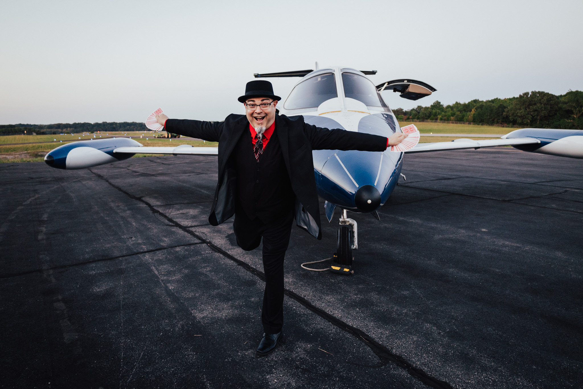 Person in black suit and hat standing in front of a small jet aircraft with arms outstretched on a runway.