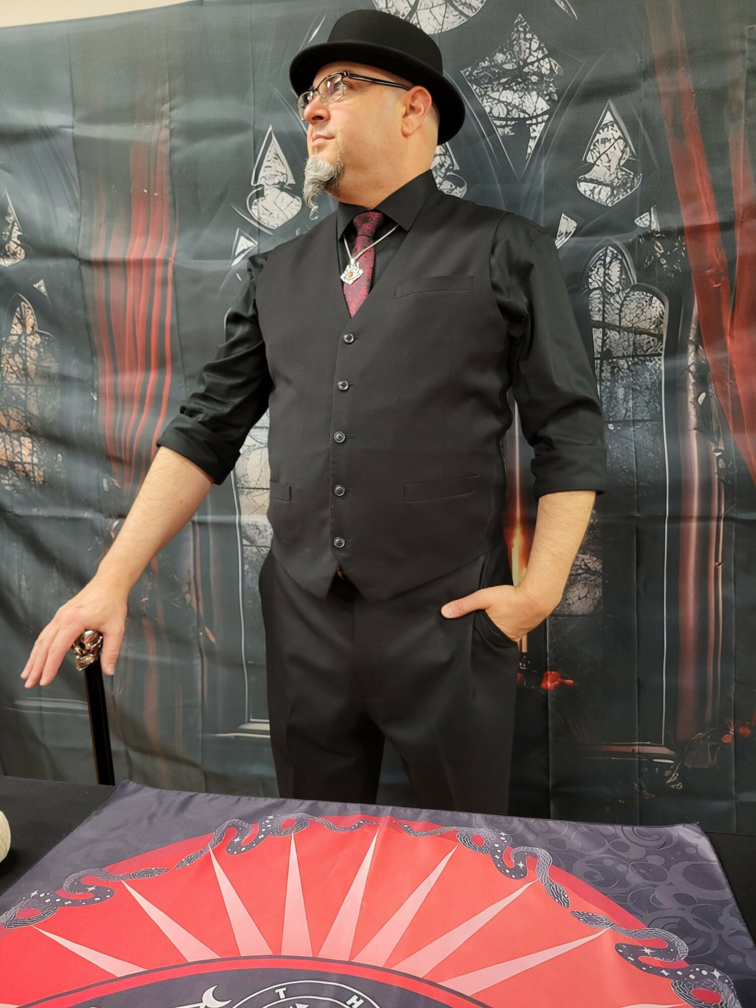 Person in black attire standing next to a table with a red and black design, holding a phone, in front of a dark backdrop.
