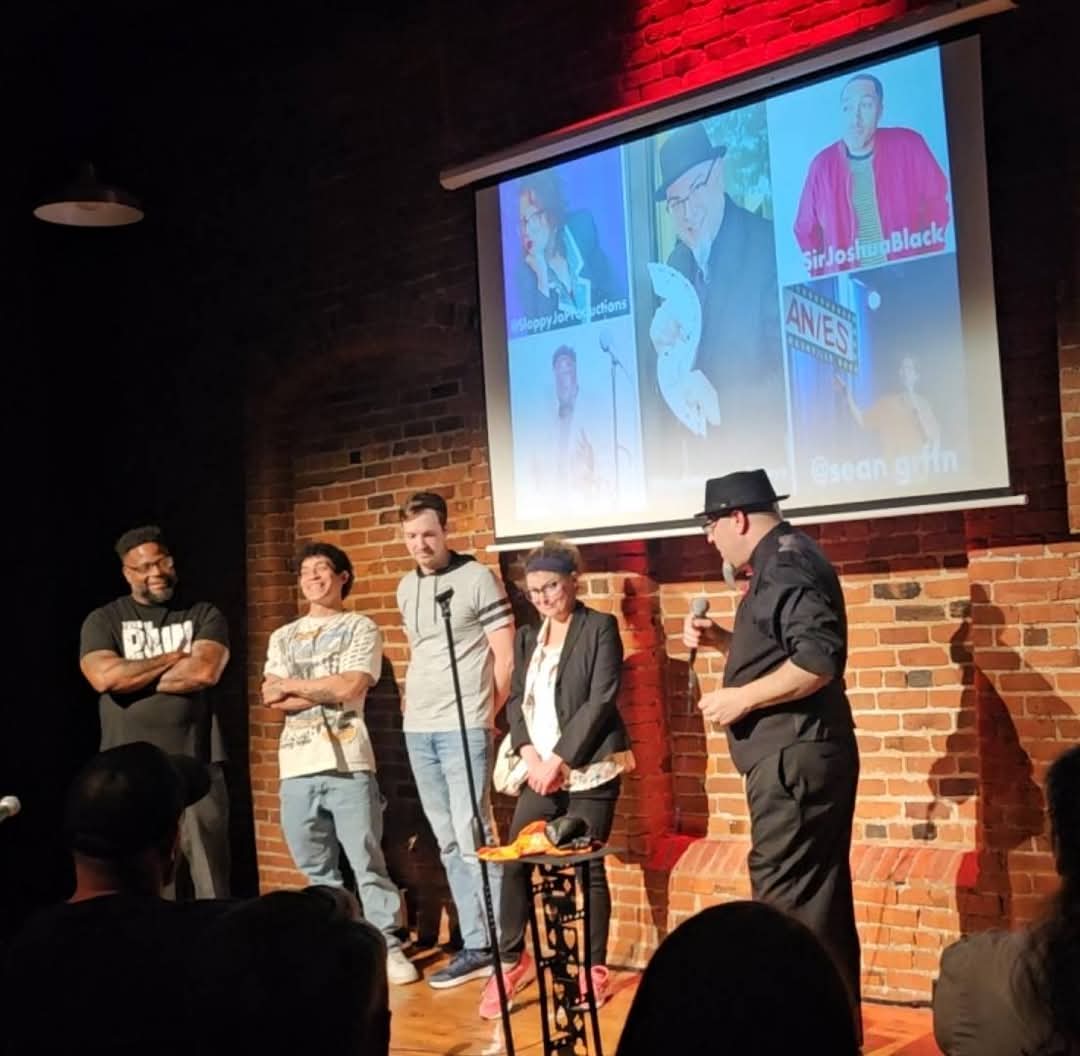 Group of five people on stage, one speaking into microphone, brick wall background, projected images above, audience in foreground.