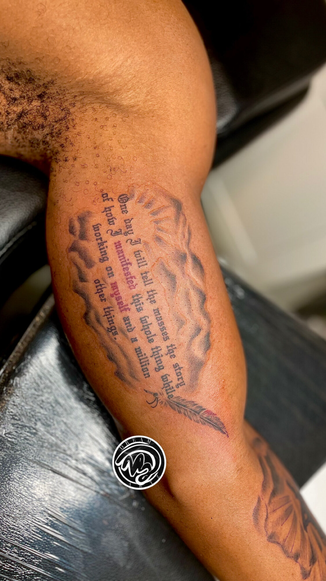 Tattoo of text and a feather on person's arm, with a dark background and part of a black shirt visible.