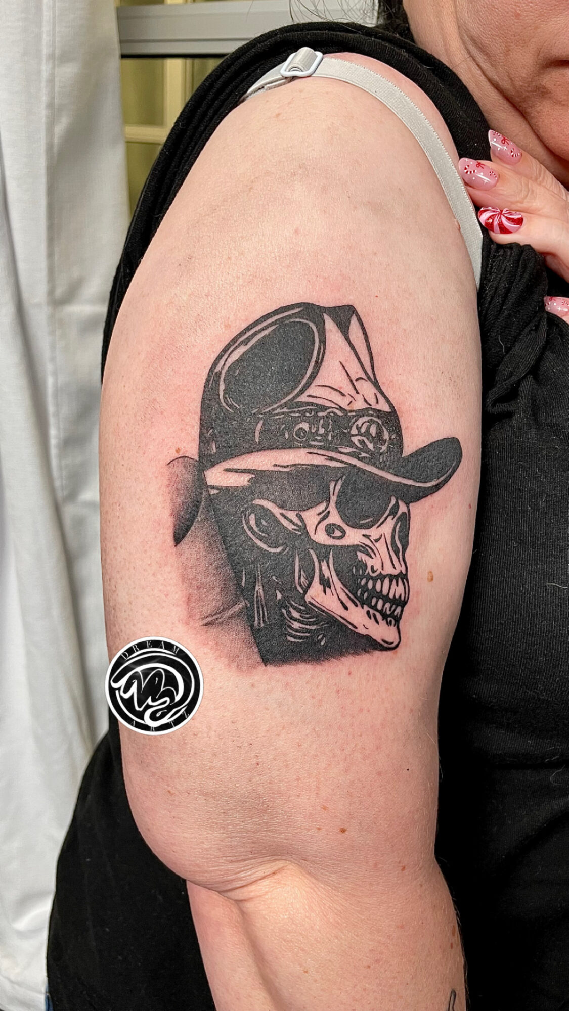 Tattoo of a skull wearing a hat on person's upper arm, with a small circular design below it.