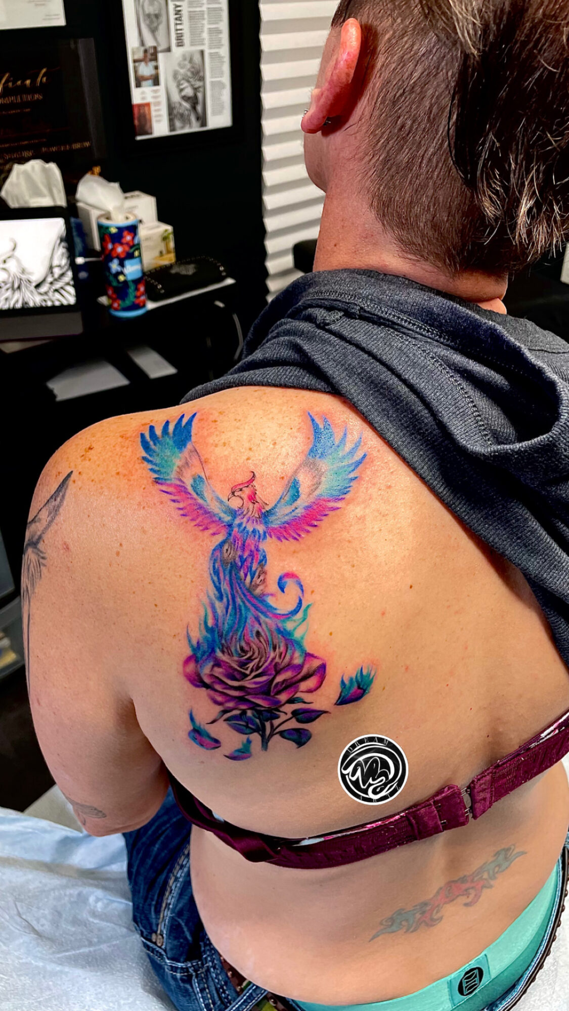 Person with a colorful phoenix tattoo on shoulder and upper back, sitting in a room with a desk and various items.