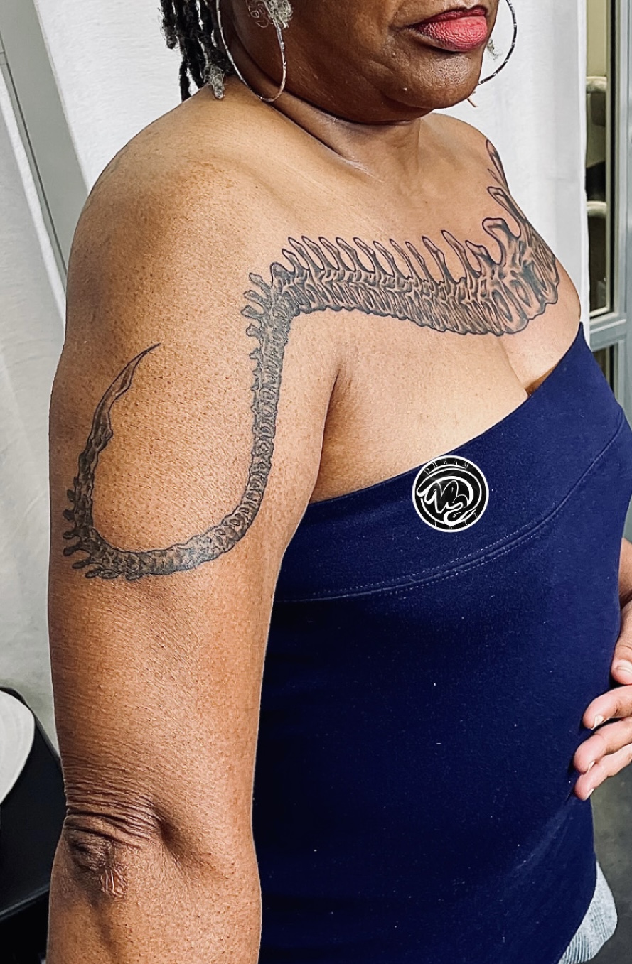 Close-up of a woman with a large snake tattoo on her shoulder and upper arm, wearing a dark blue strapless top.
