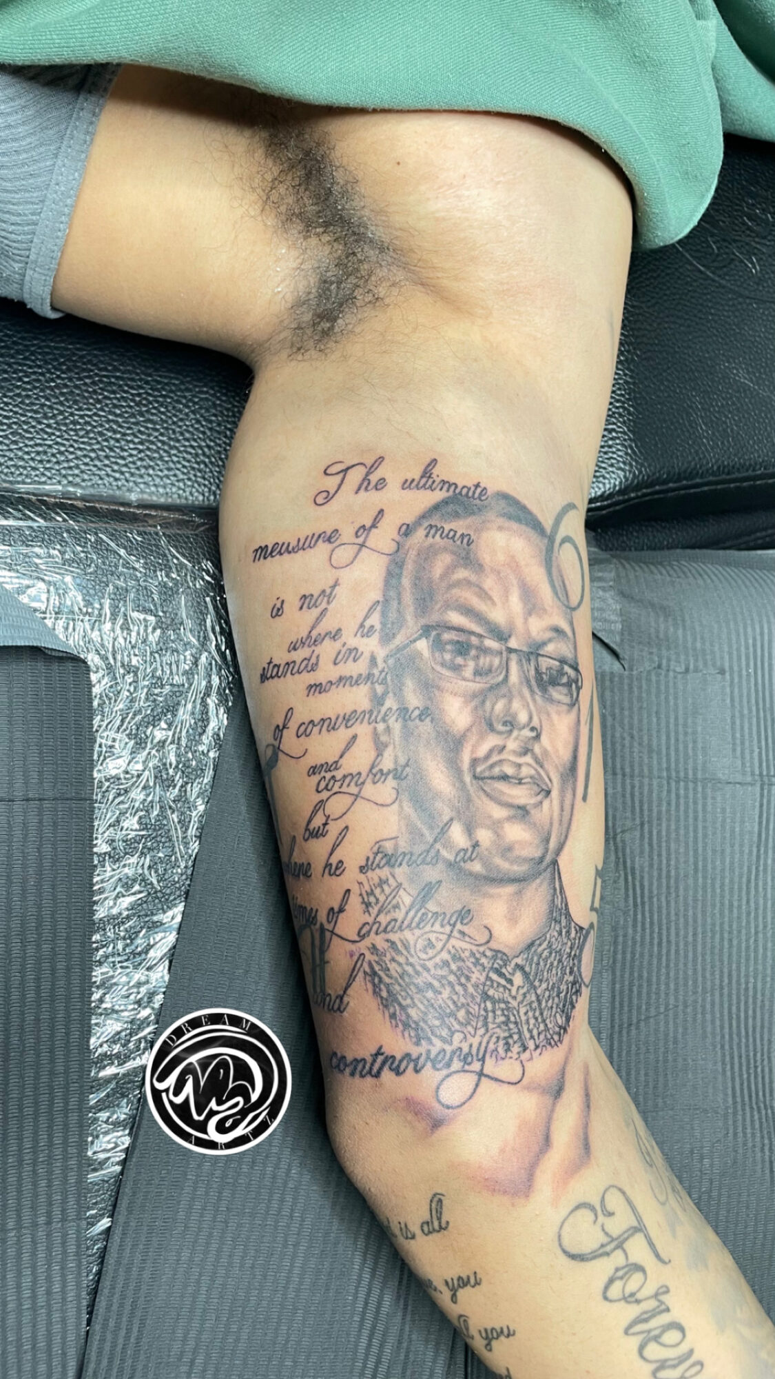 Tattooed arm with a portrait of a man wearing glasses, surrounded by cursive text, resting on a surface with a green sleeve visible.