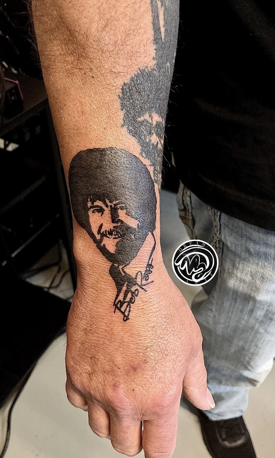 Tattoo of a smiling person's face with dark hair on a forearm, with smoke or cloud design above, and a signature below.