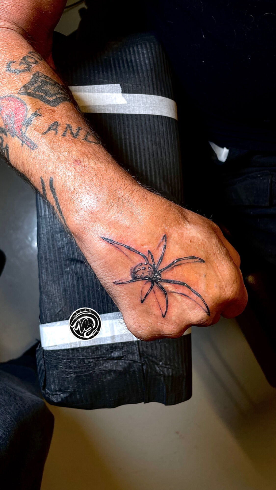 Tattooed arm with a spider tattoo on the hand, resting on a black object, with other tattoos visible on the arm.