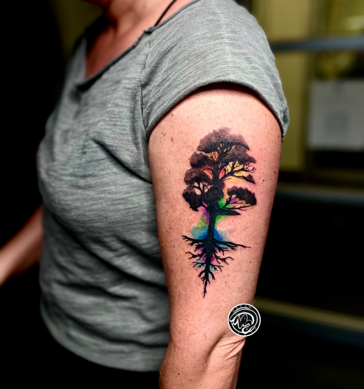 Person with a colorful tree tattoo on upper arm, showing roots and branches, wearing a gray shirt.