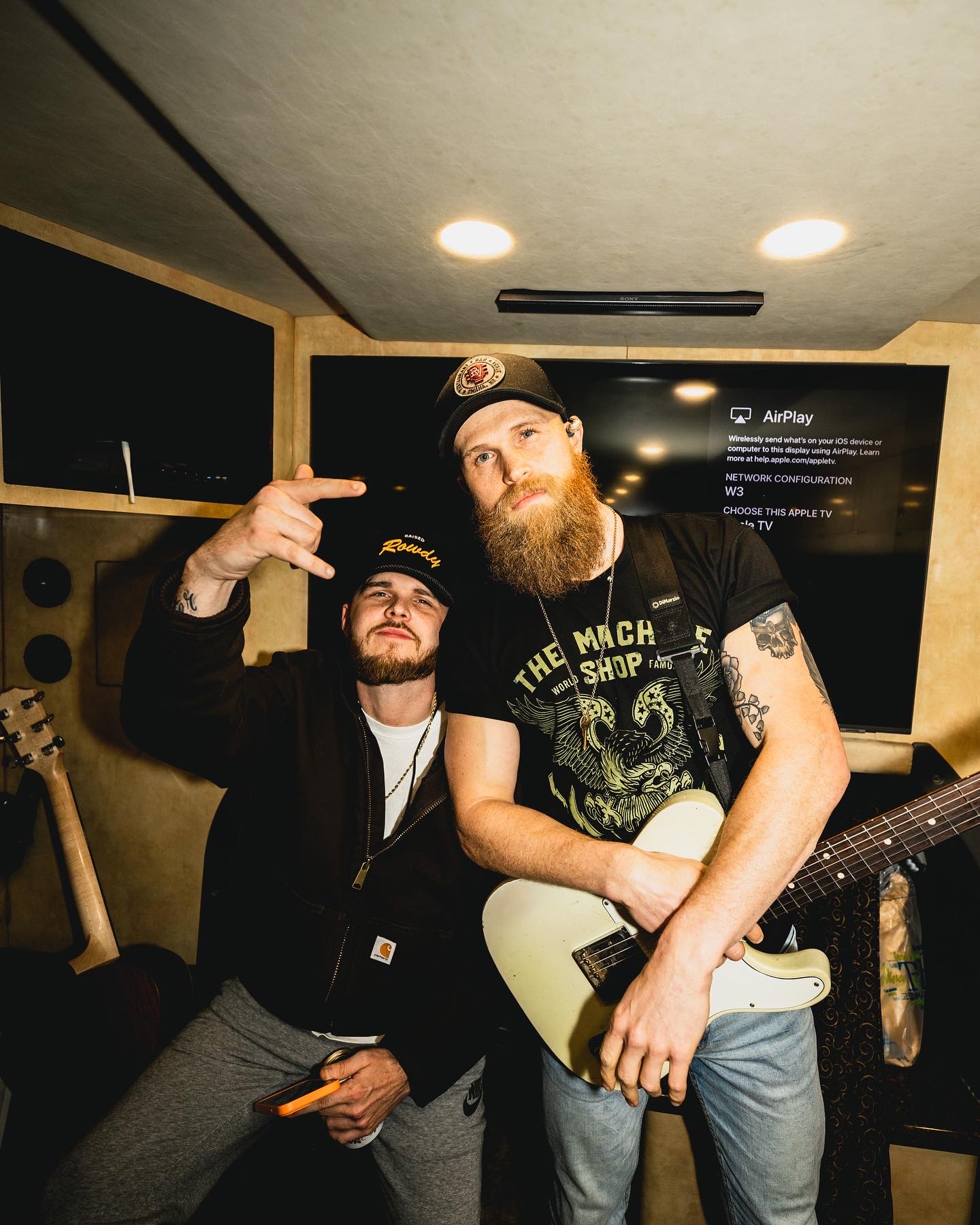 Two men in a music studio, one with a guitar, the other making a gesture, both wearing casual clothing and hats.