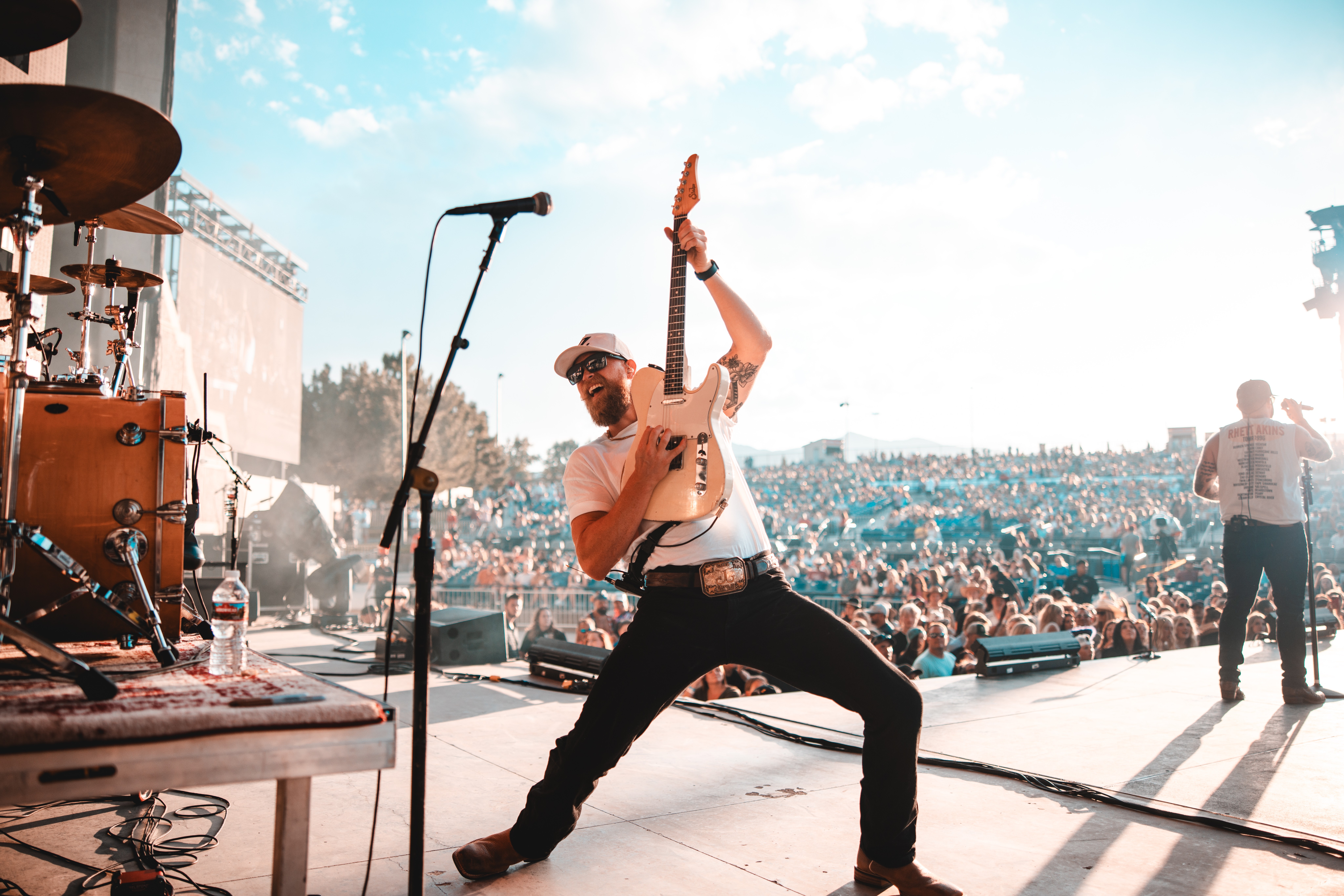 Musician on stage playing guitar with a large crowd in the background, bright sky, and stage equipment visible.