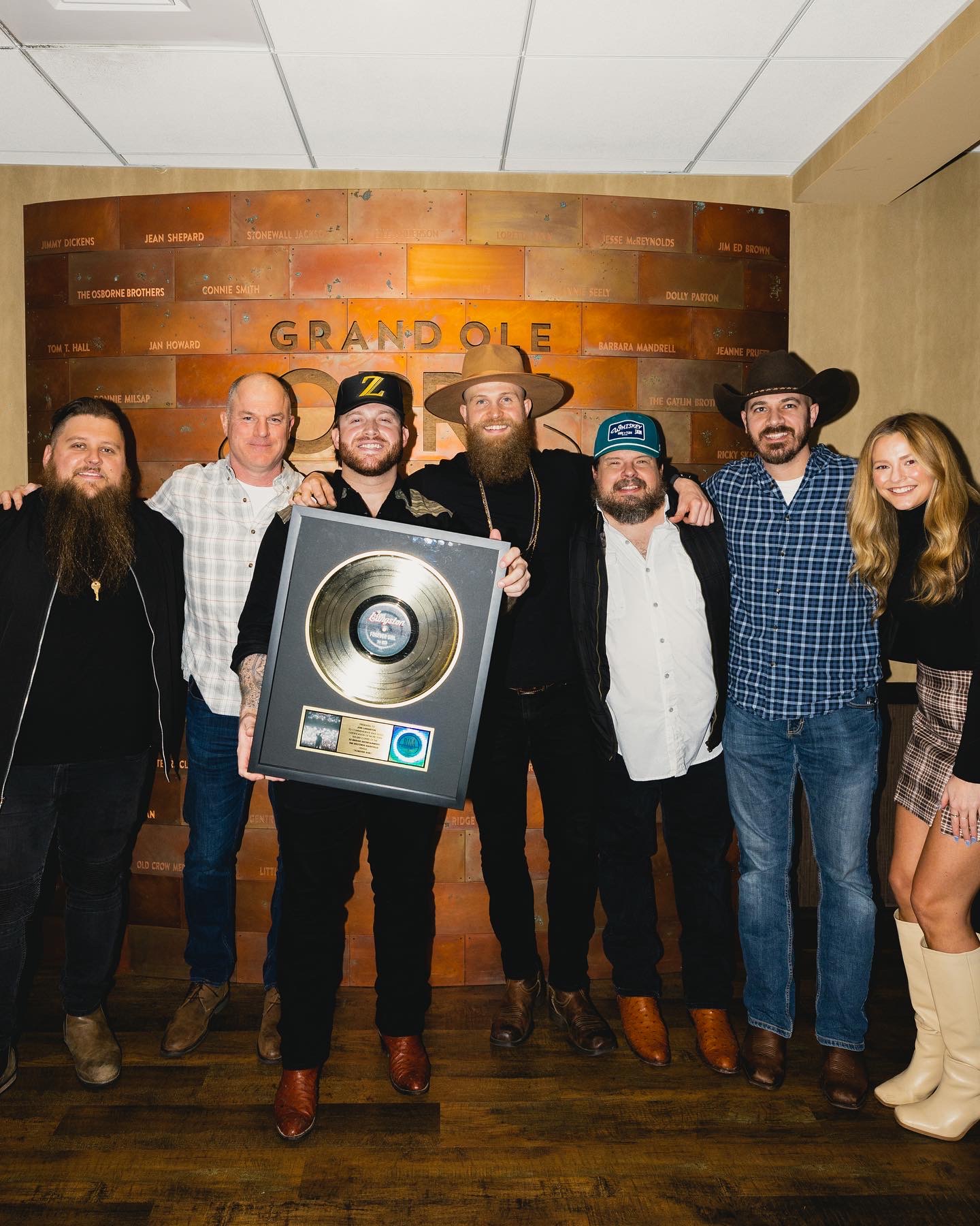 Group of seven people standing in front of a brick wall with a sign that says 'Grand Ole Oe', one person holding a gold record plaque, all smiling.