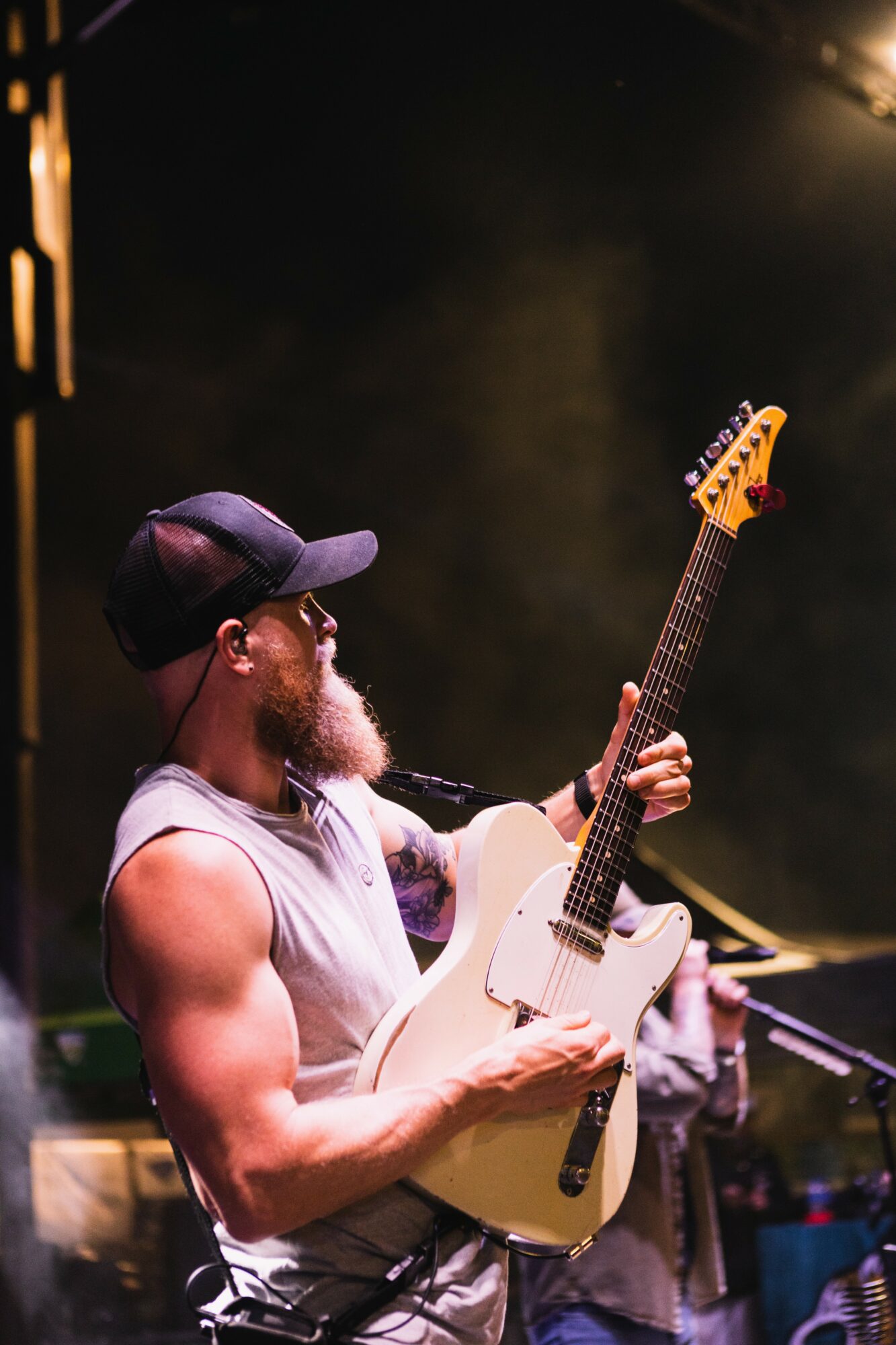 Musician with a beard, wearing a cap and sleeveless shirt, playing an electric guitar on stage with dark background.