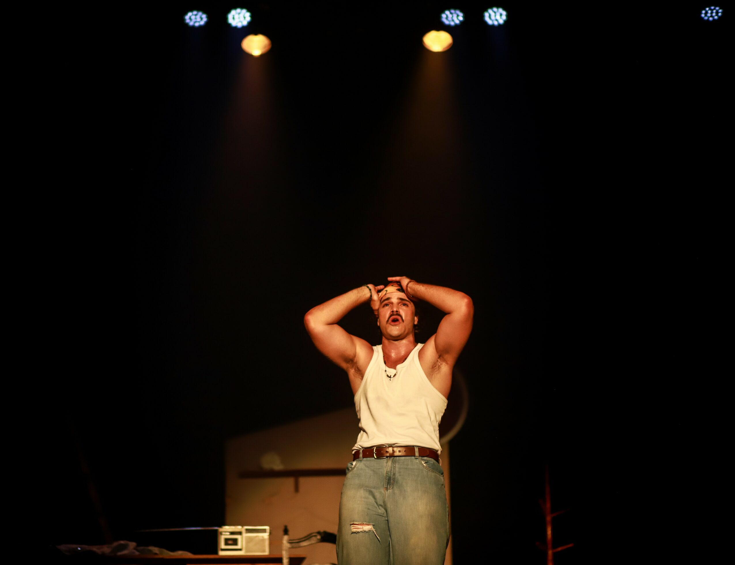 Person on stage with hands on head, illuminated by stage lights, dark background, wearing a white tank top and jeans.