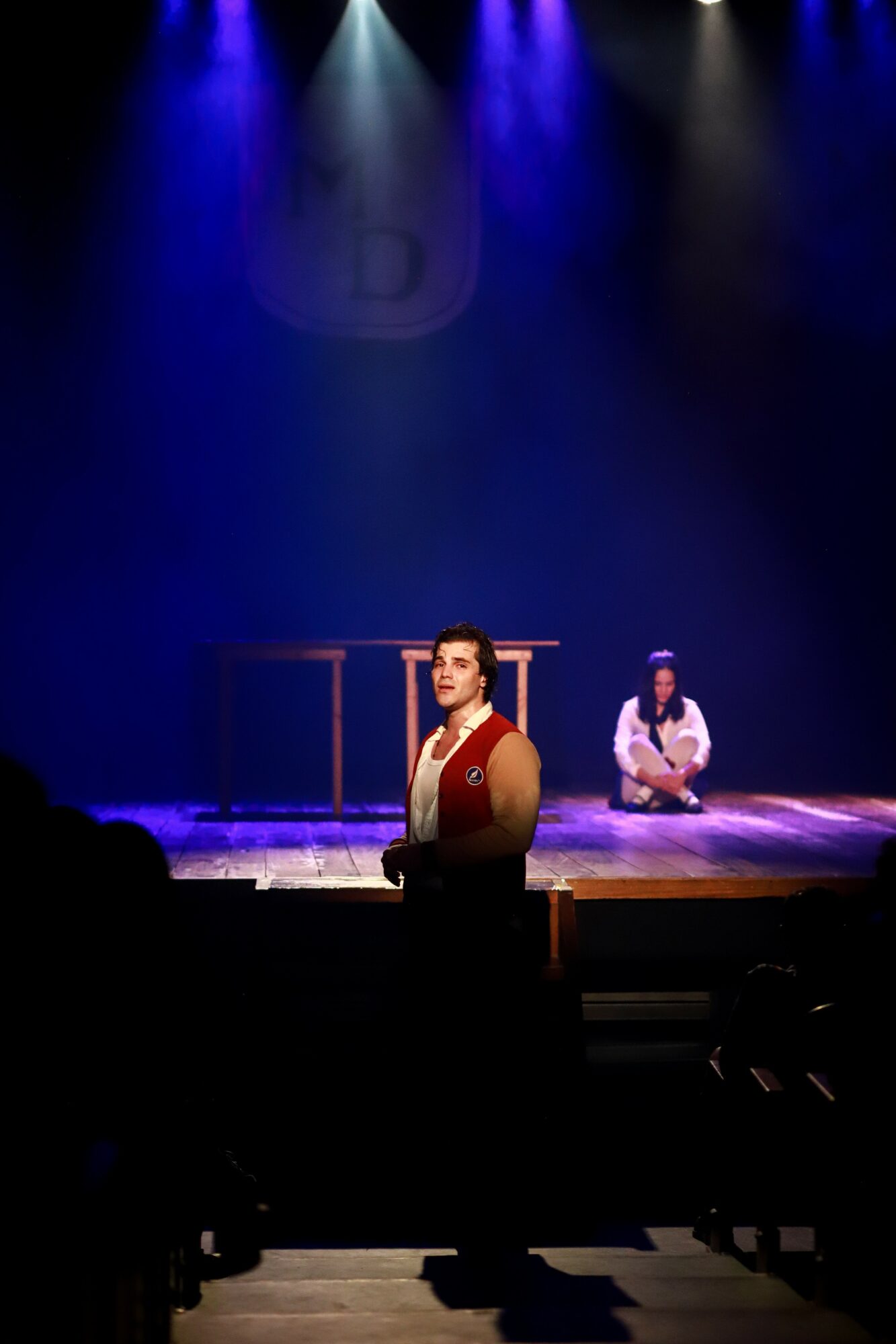 Two performers on stage, one standing in front and one sitting in the background, with blue and purple stage lighting.