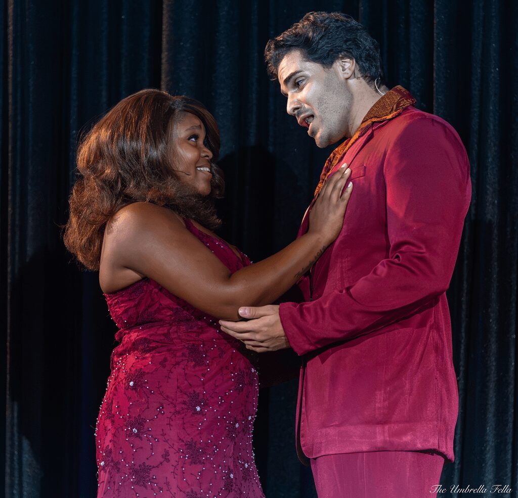 Two people, a woman and a man, face each other with the woman touching the man's chest, both smiling, against a dark curtain background.