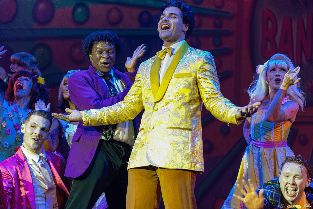 Group of performers on stage, smiling and singing, colorful costumes, lively atmosphere.