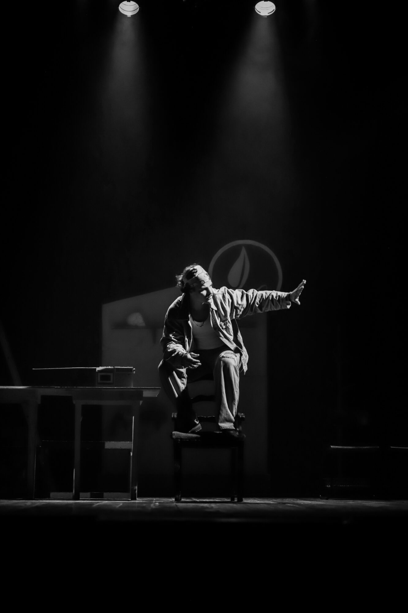 Person sitting on a chair on stage, pointing with one arm, illuminated by stage lights, in black and white.