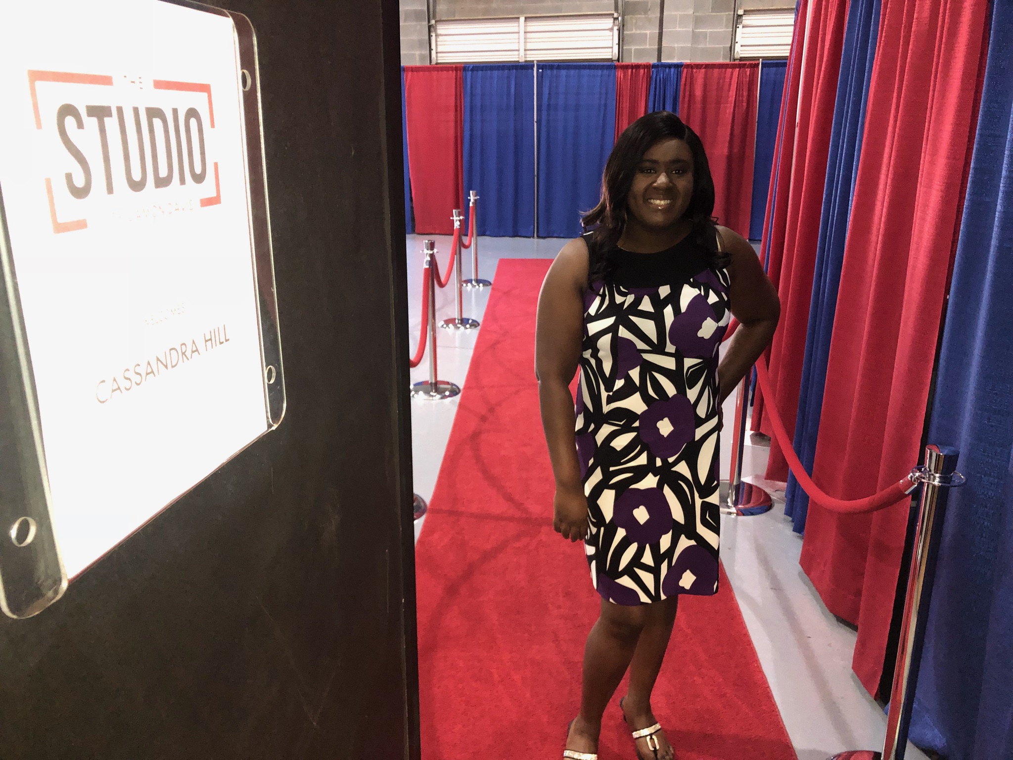 Woman standing on a red carpet with red and blue curtains in the background, smiling, wearing a patterned dress.