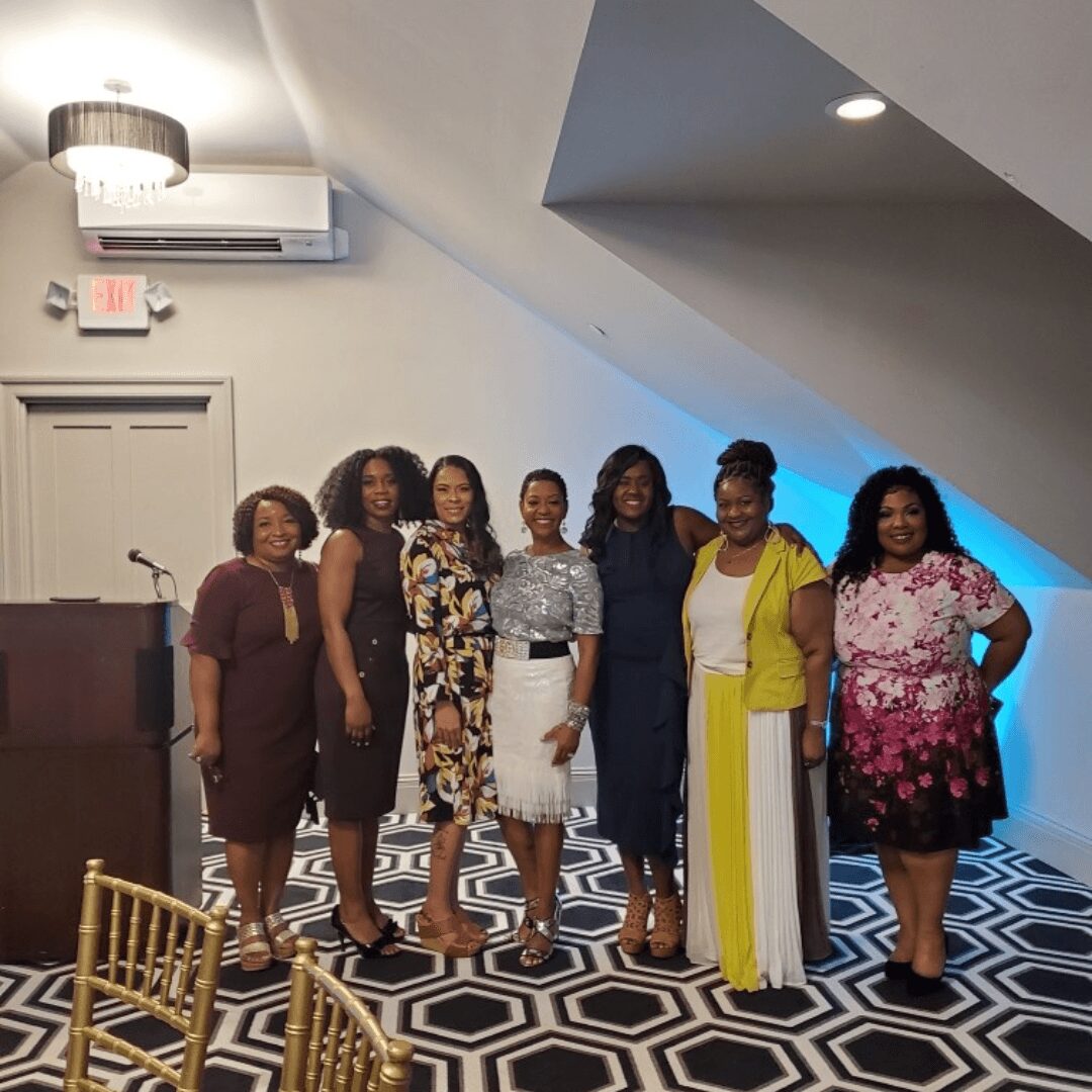 Group of seven women standing together indoors, smiling, with patterned floor and white walls, some wearing colorful dresses.
