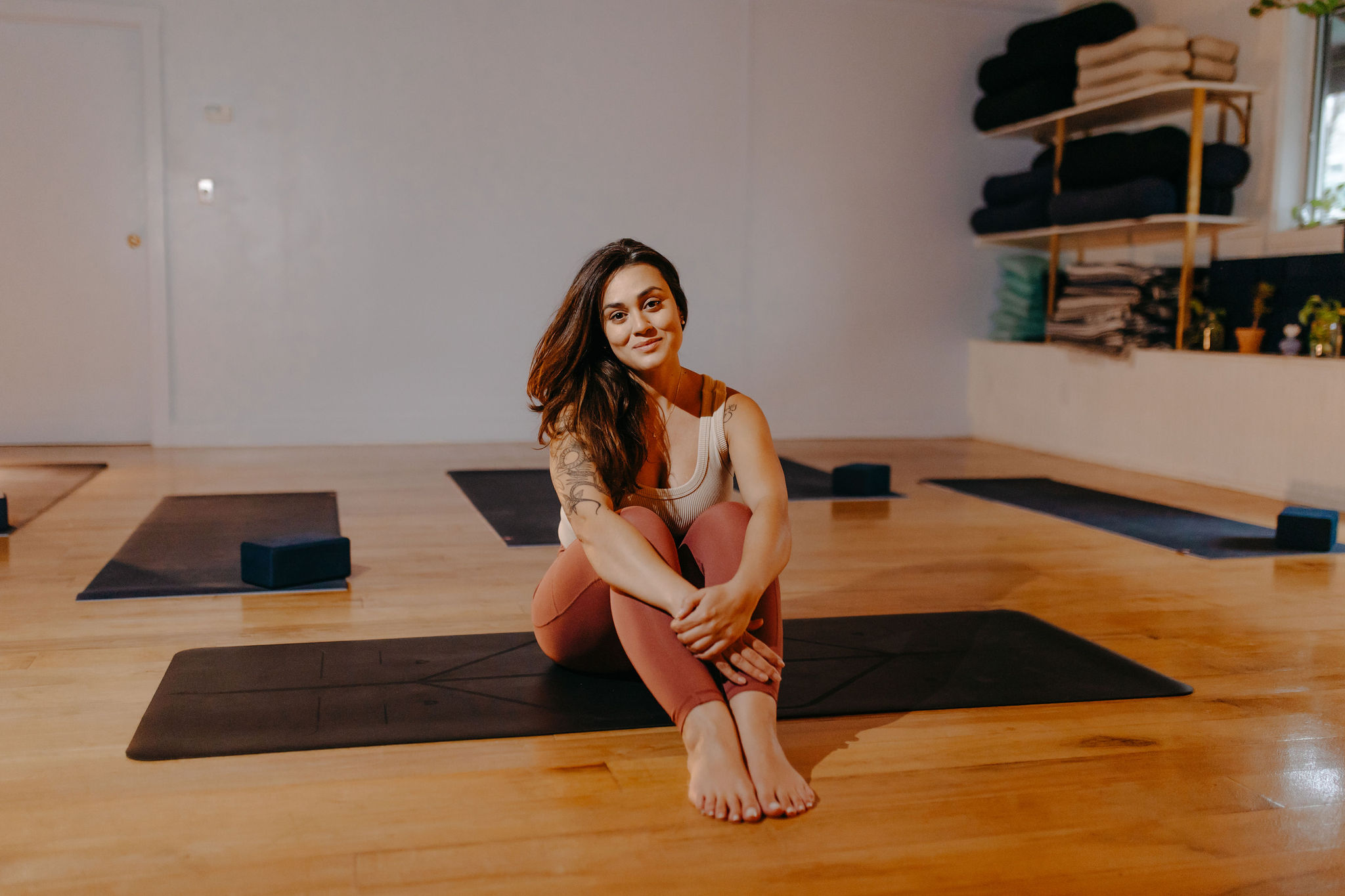 Woman sitting on yoga mat in a studio with shelves of folded towels and yoga props, smiling at the camera.