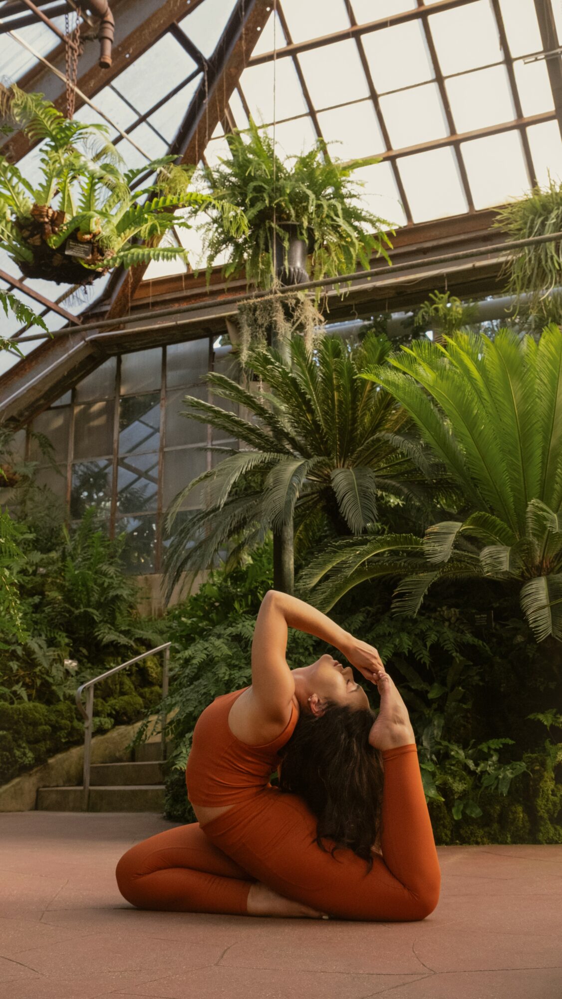 Person in orange outfit performing a yoga pose in a greenhouse with lush green plants and glass ceiling.