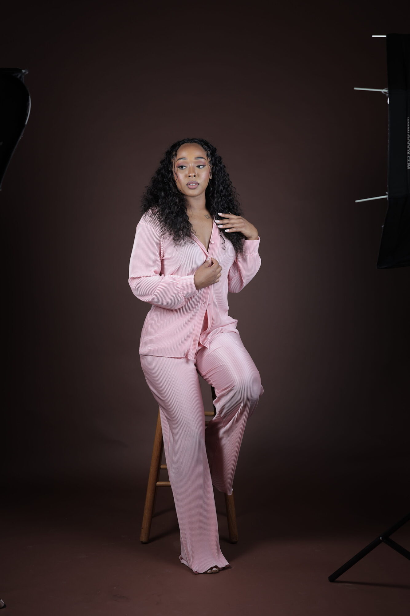 Woman with curly hair in pink pajamas sitting on a stool against a dark background, studio lighting setup visible.