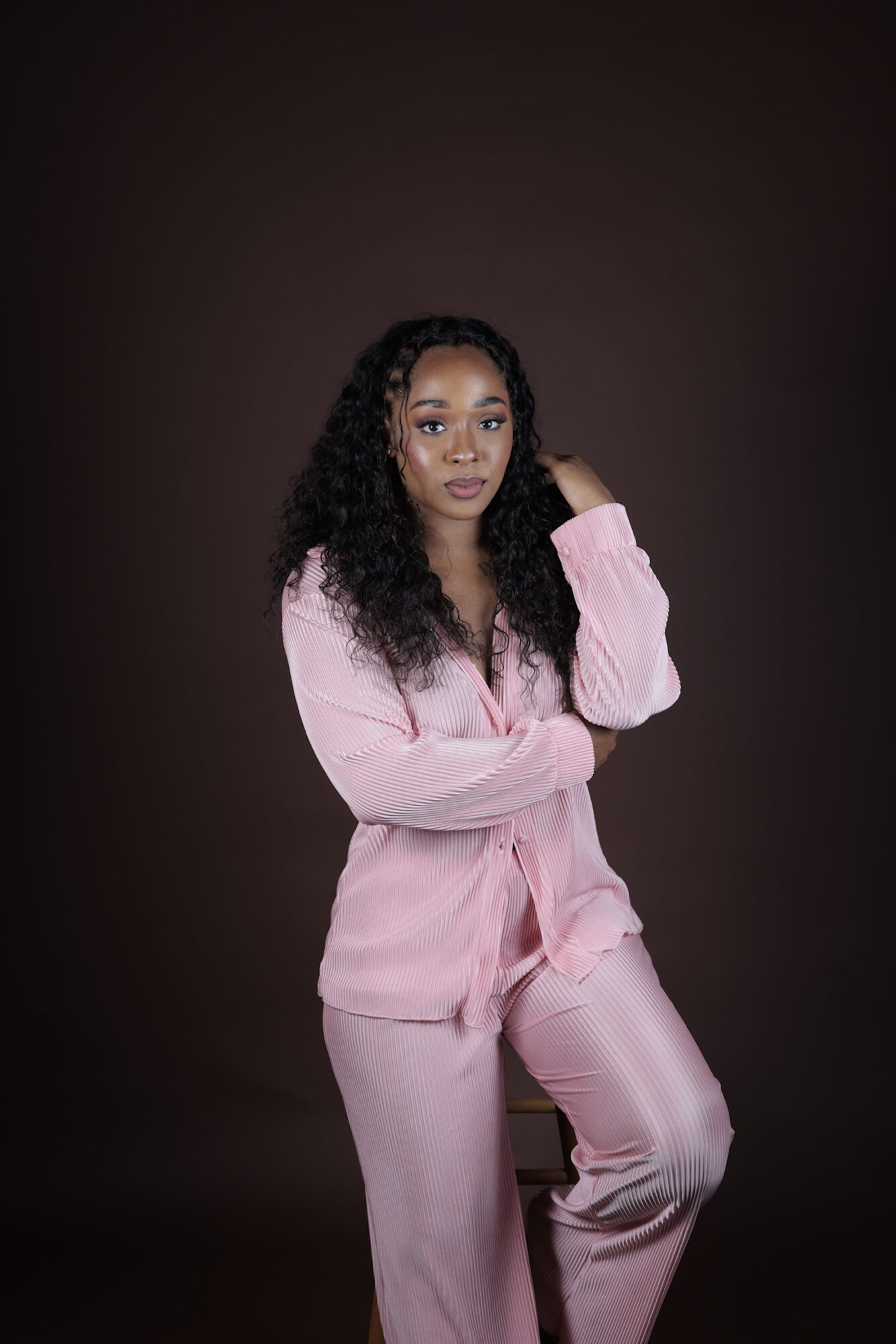 Young woman with curly hair wearing pink pajamas, standing against a dark background, looking at the camera with a neutral expression.