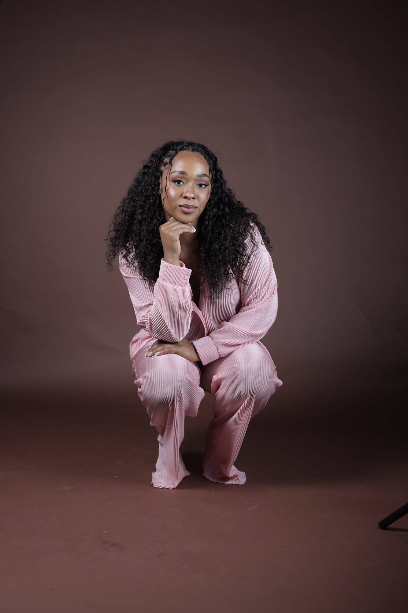 Woman with curly hair in pink pajamas crouching against a brown background.