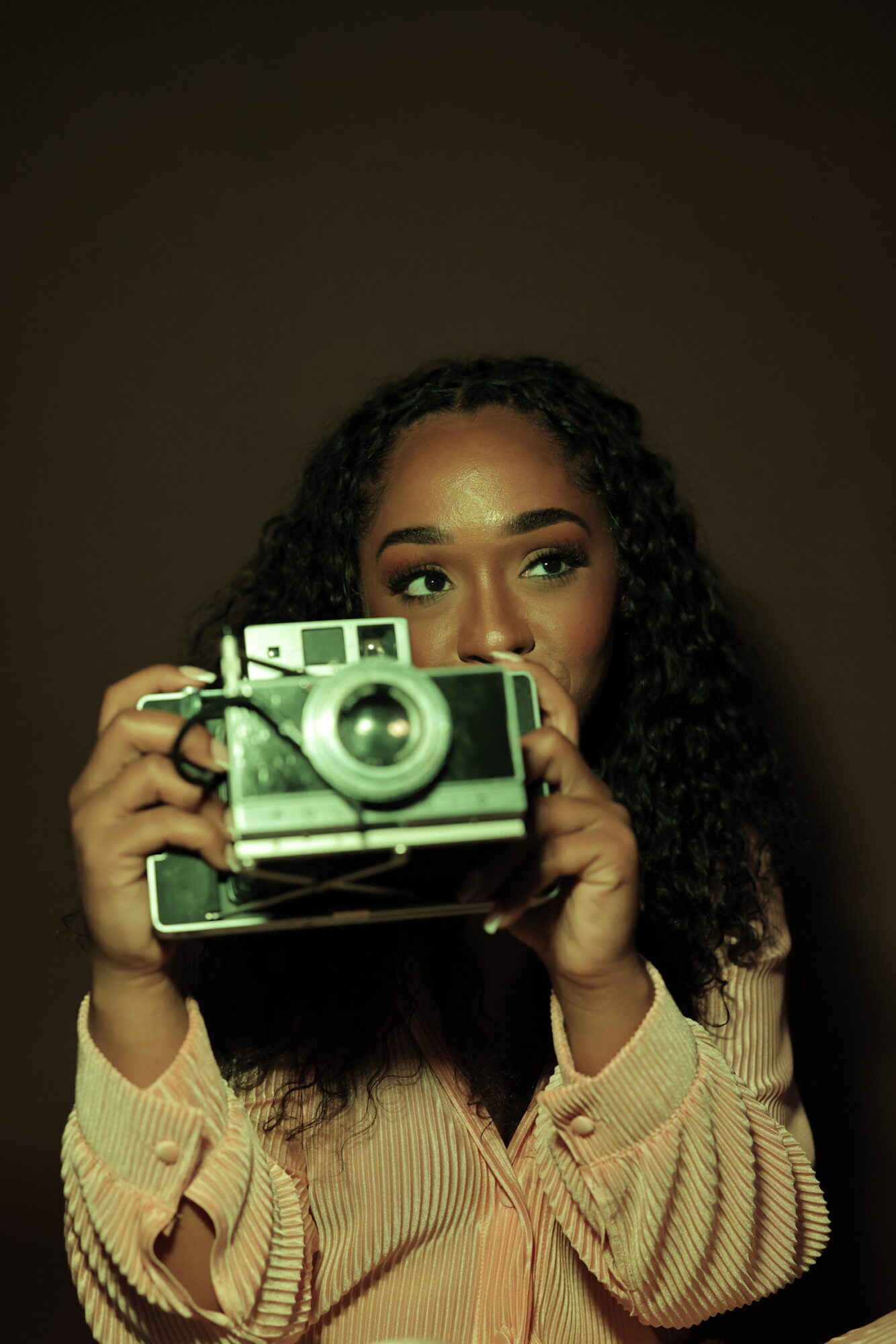 Young woman with curly hair holding a vintage camera, partially covering her face, against a dark background.