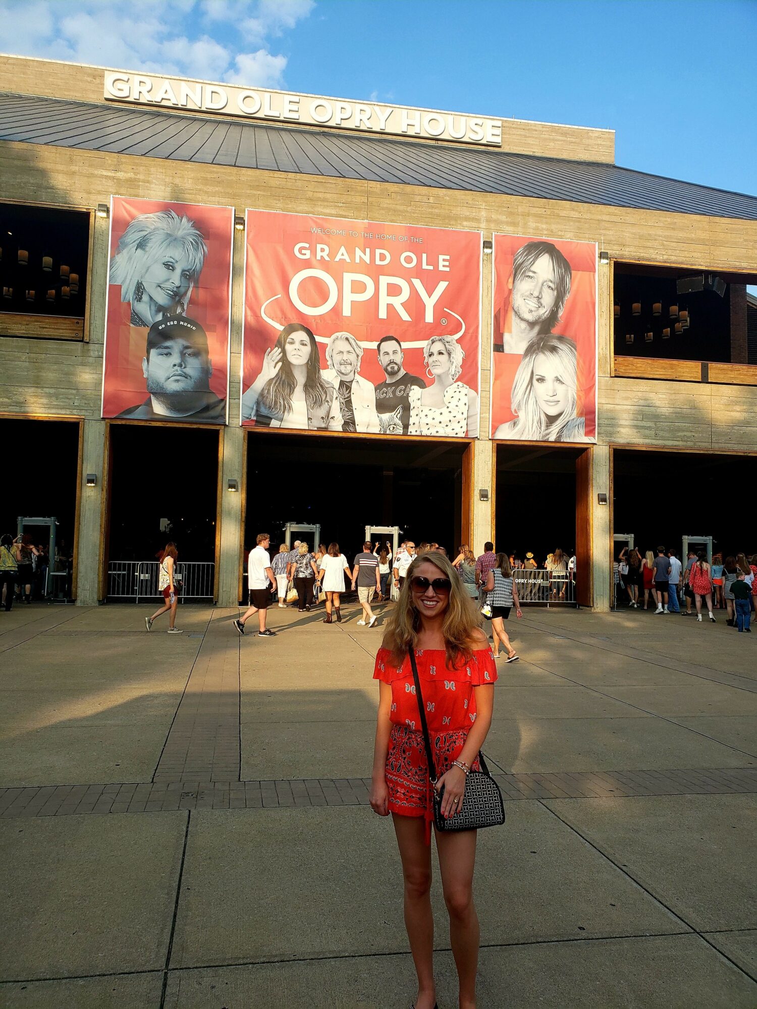 Woman in red dress and sunglasses standing in front of Grand Ole Opry building with banners, people, and blue sky.