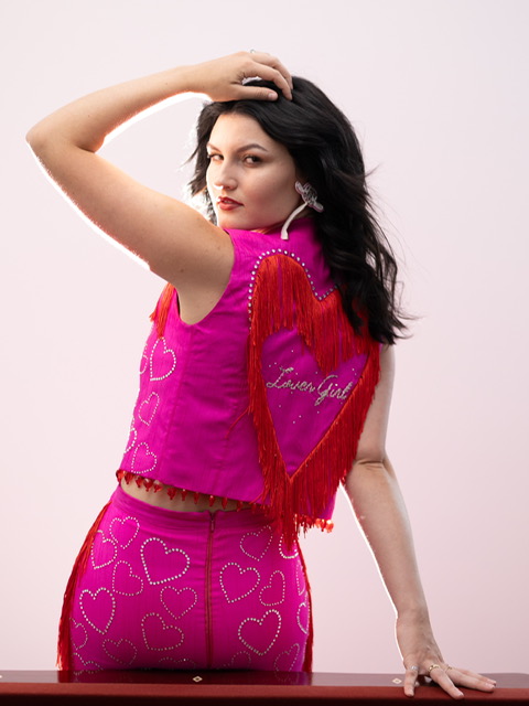 Woman with dark hair in a pink heart-patterned outfit, posing with one hand on her head and the other on a surface.