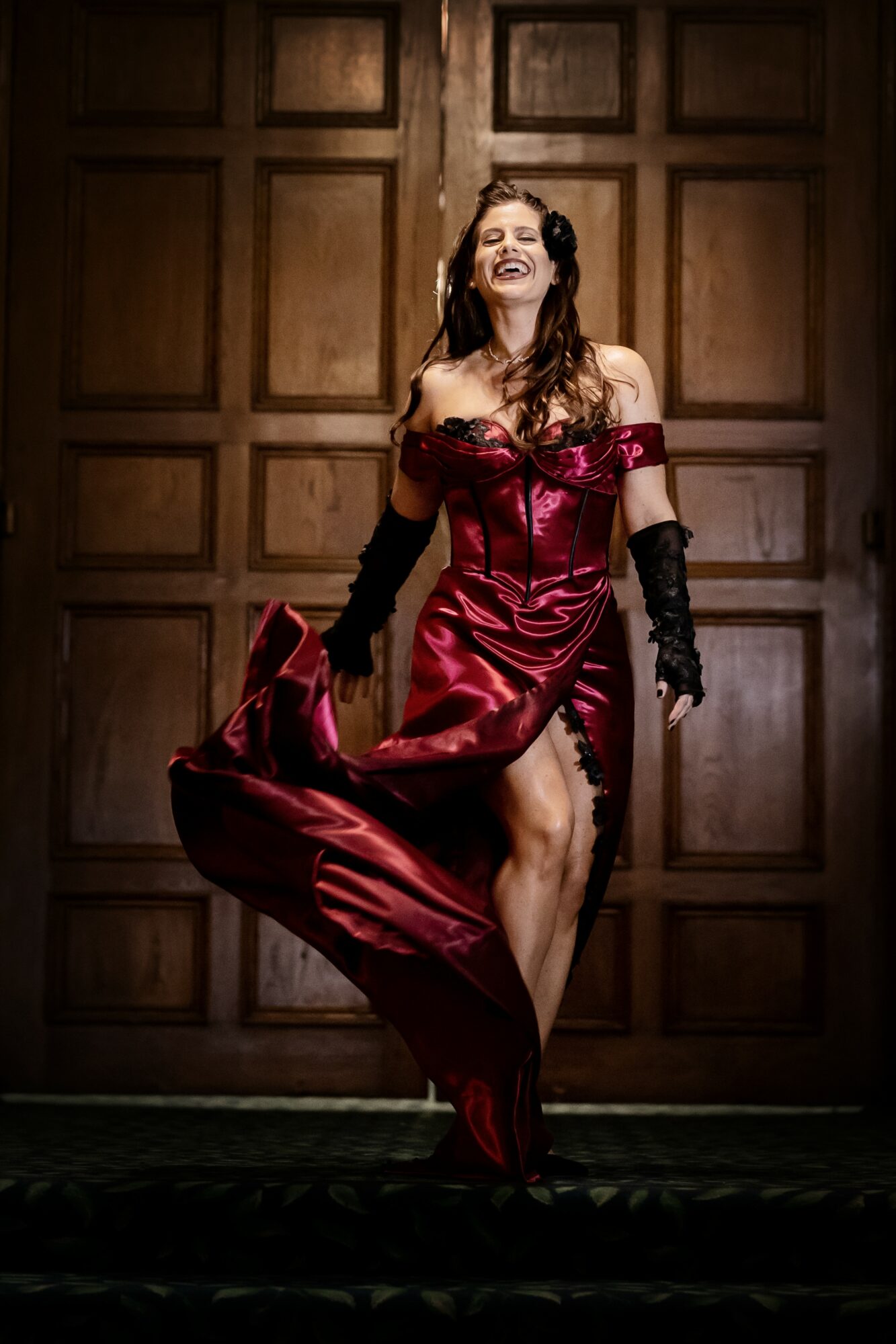 Woman in a red dress lifting her skirt, smiling, in front of wooden panel background.