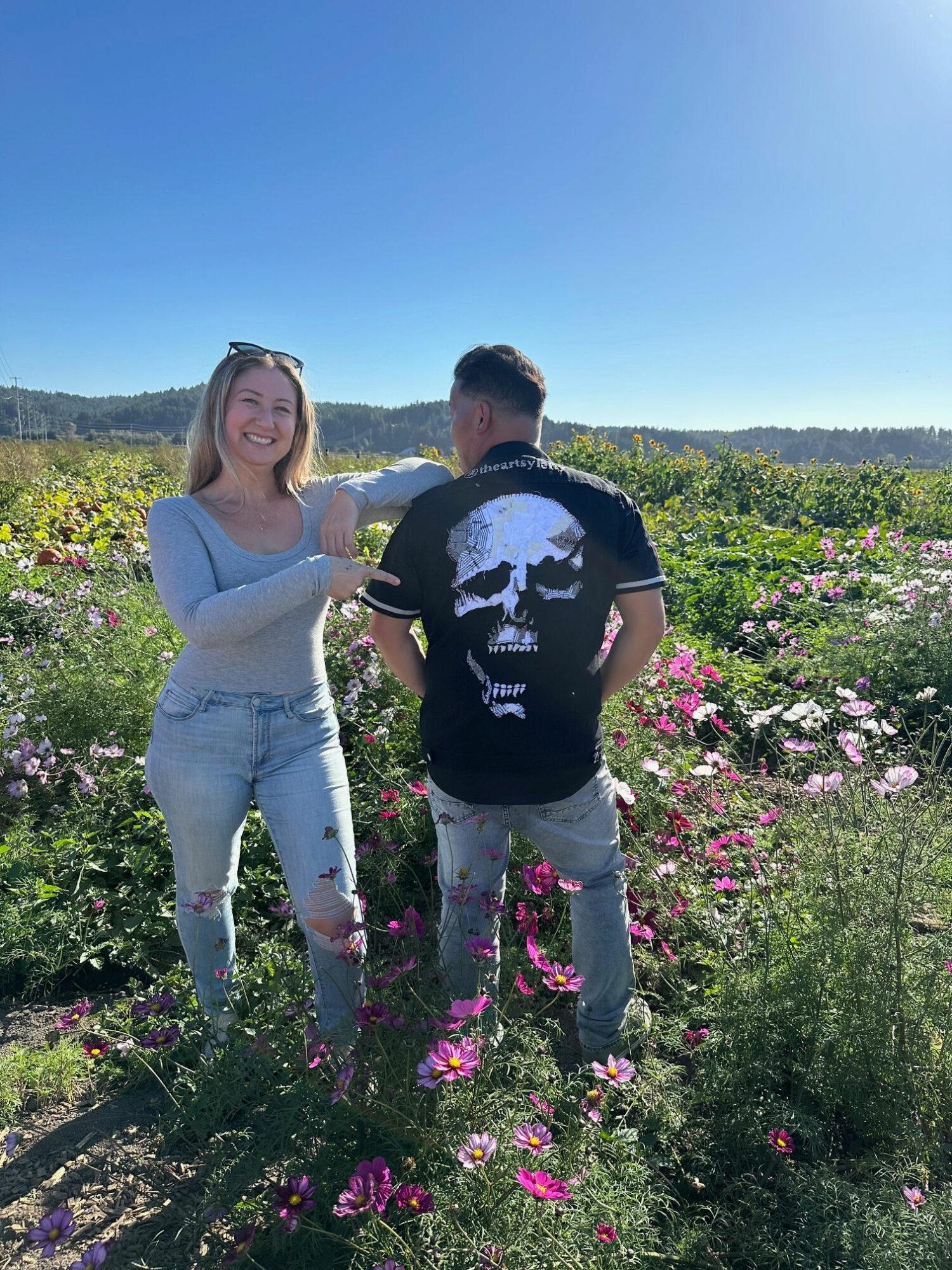 A man and woman stand in a flower field with a blue sky and distant landscape, holding hands and smiling.
