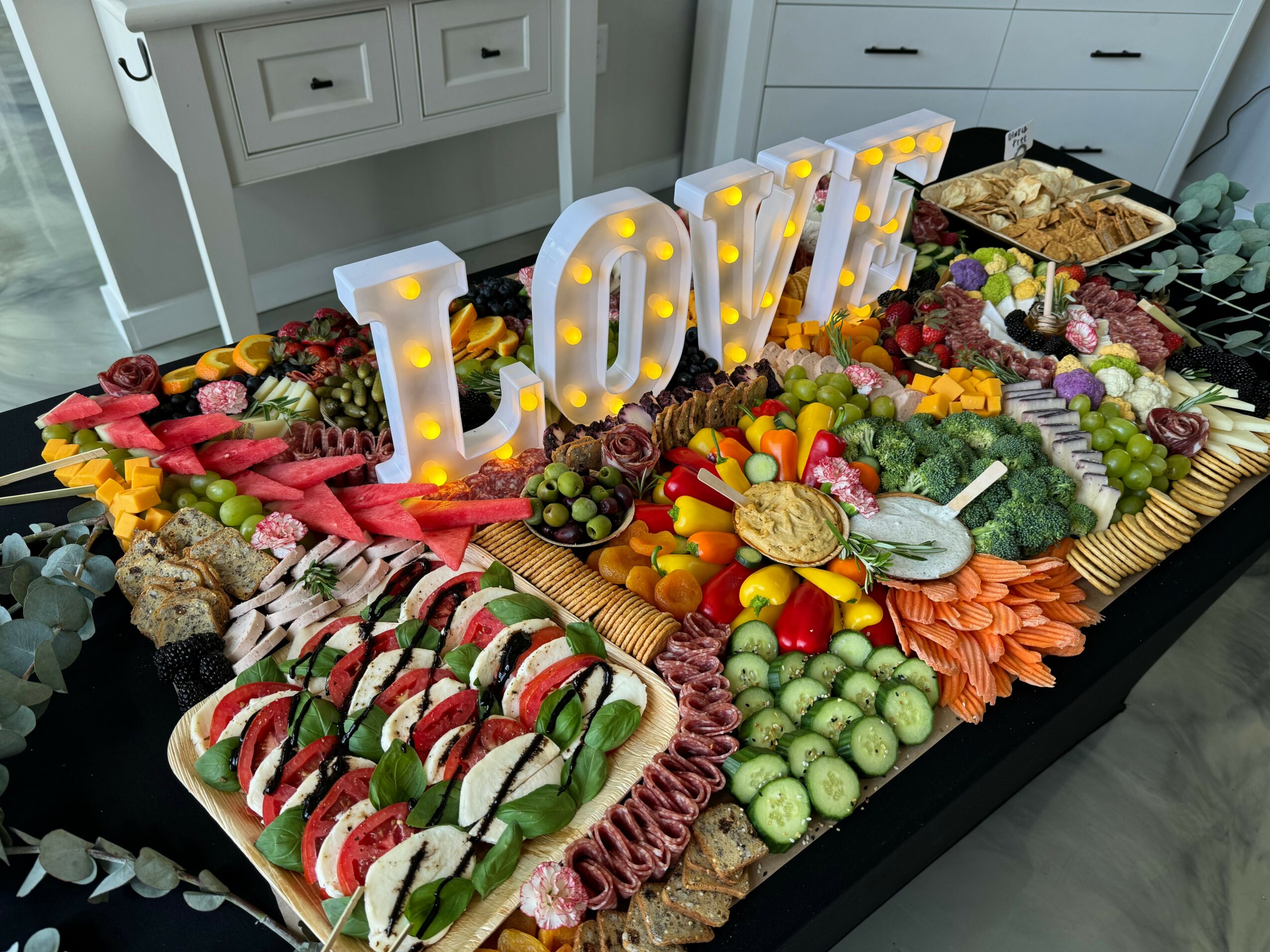 Table with a large floral arrangement, illuminated 'LOVE' sign, and a platter of sliced tomatoes and basil, in a bright room.