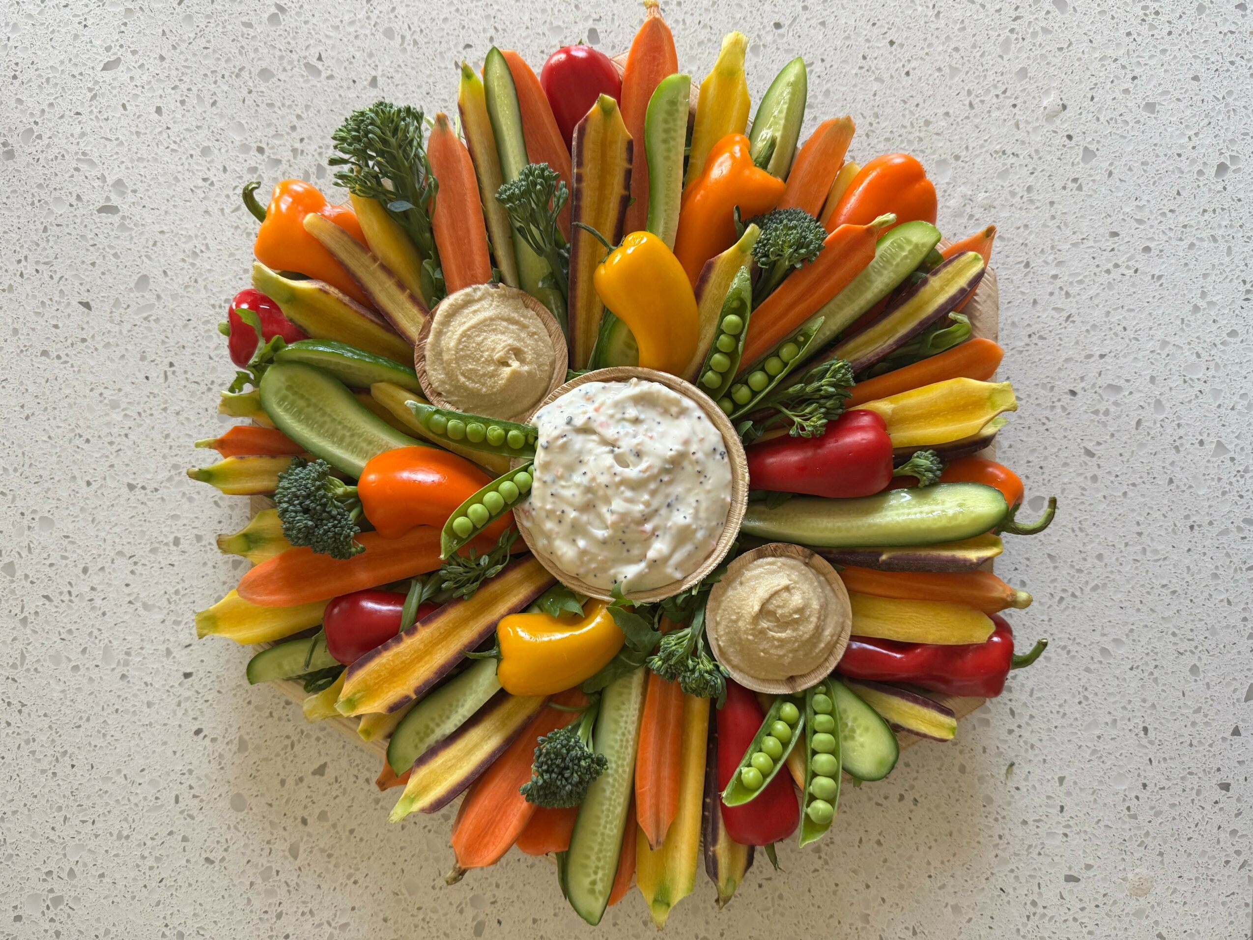 Colorful vegetable platter arranged in a circular pattern with a white dip in the center, on a speckled surface.