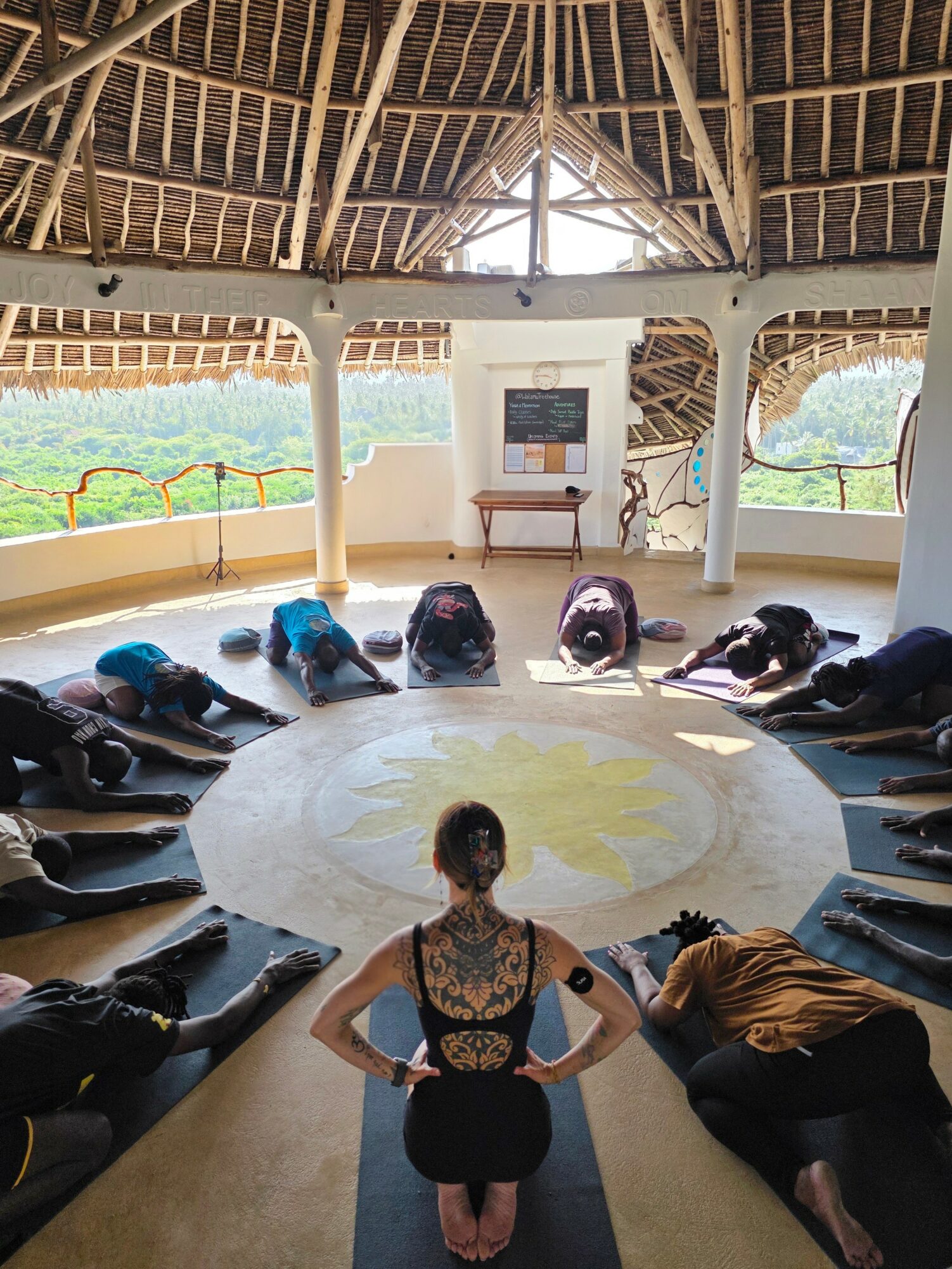 Group of people lying on the floor in a circle inside a thatched-roof room, with a woman kneeling in the center facing away.