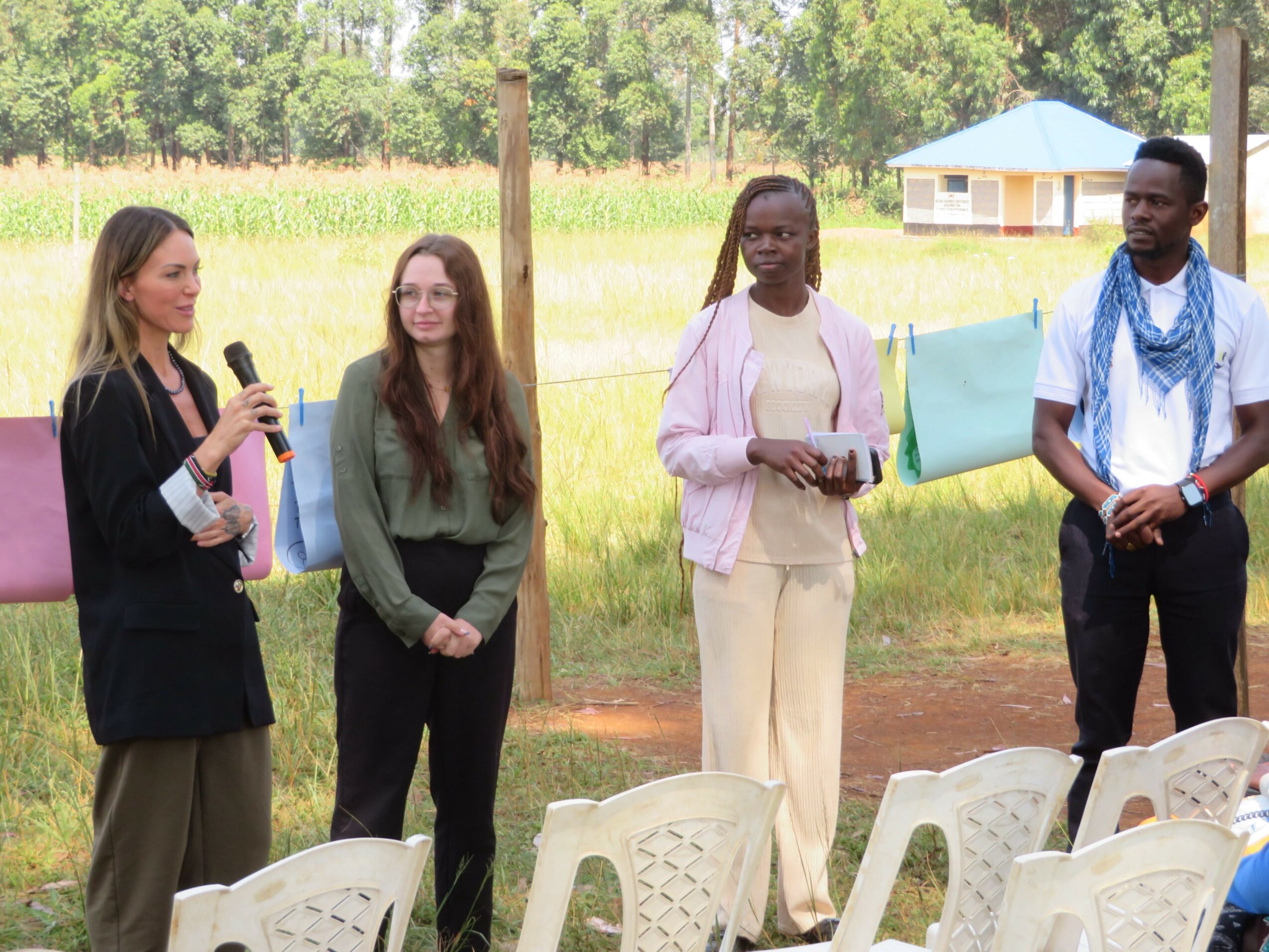 Four women standing outdoors near chairs, with a field and small building in the background.