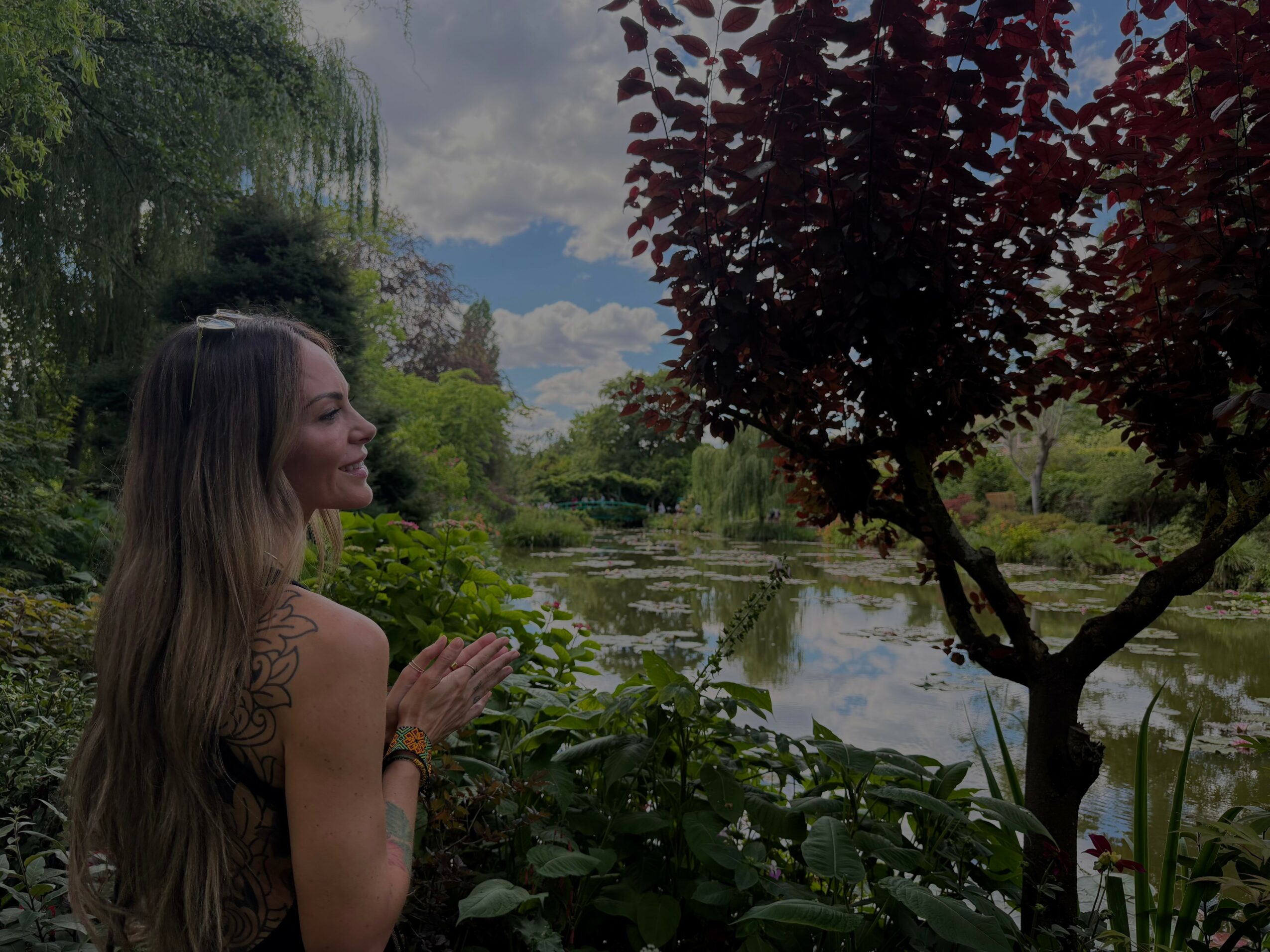 Woman with long hair smiling near a pond with trees and greenery, cloudy sky in background.