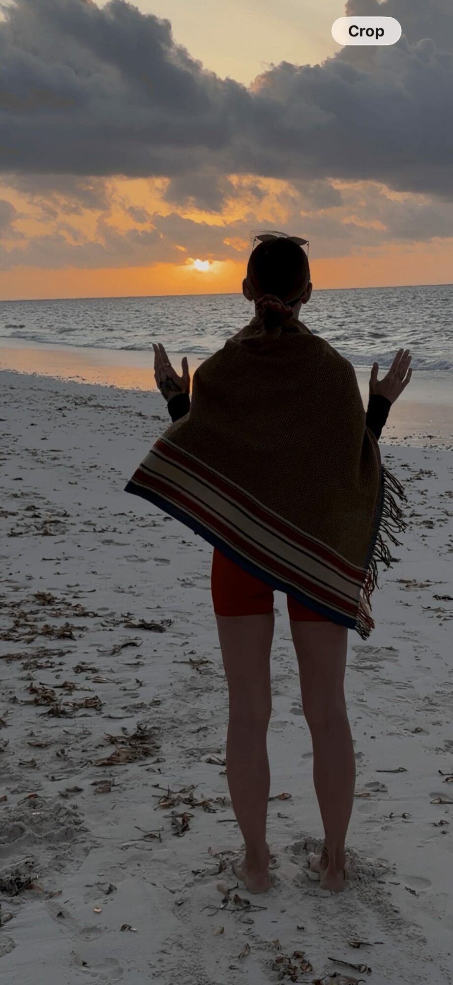 Person standing on beach facing sunset, arms outstretched, wearing a shawl and shorts, sandy shore in foreground, cloudy sky.