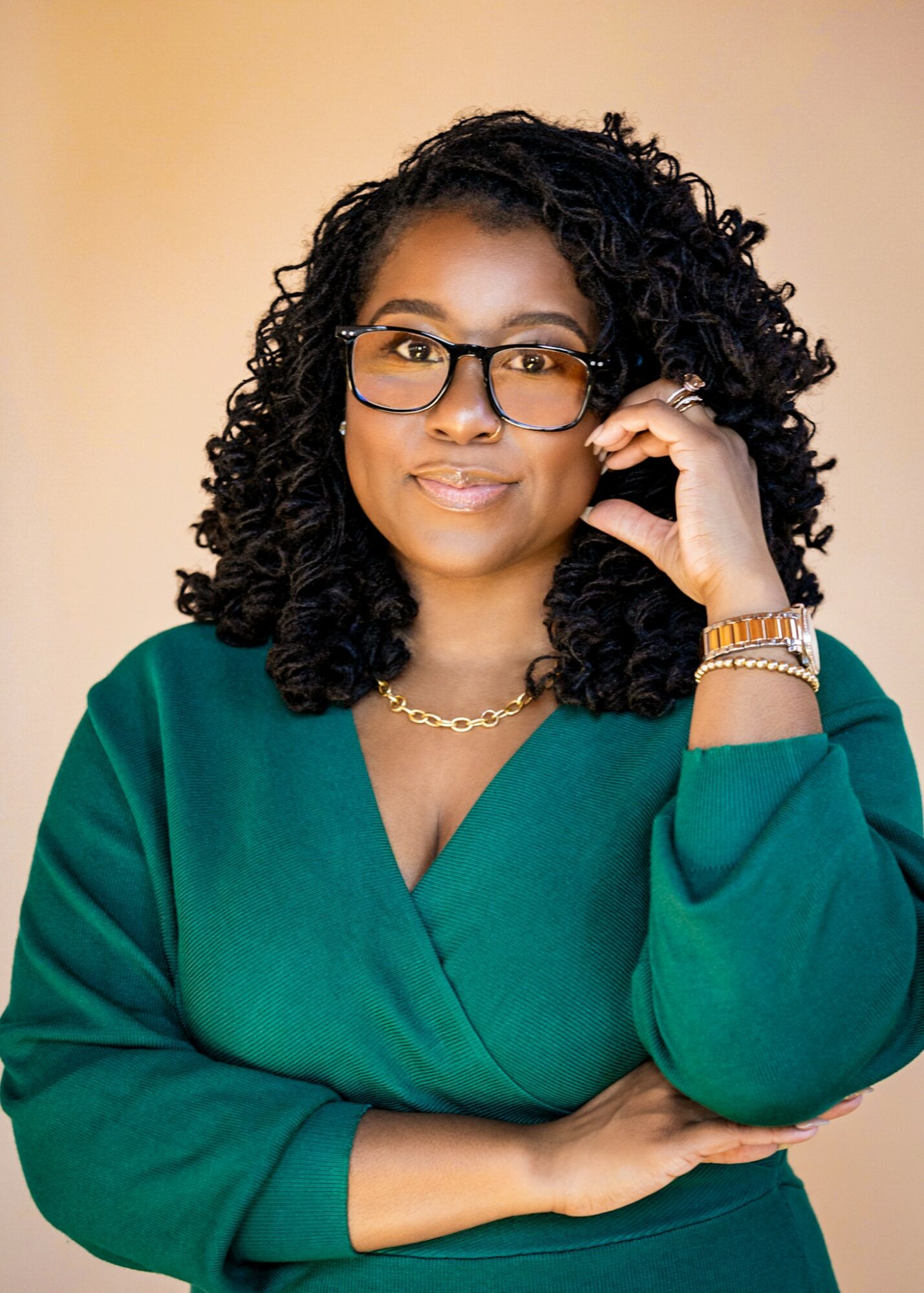 Woman with curly hair wearing glasses, a green top, and jewelry, posing with hand near face against a neutral background.