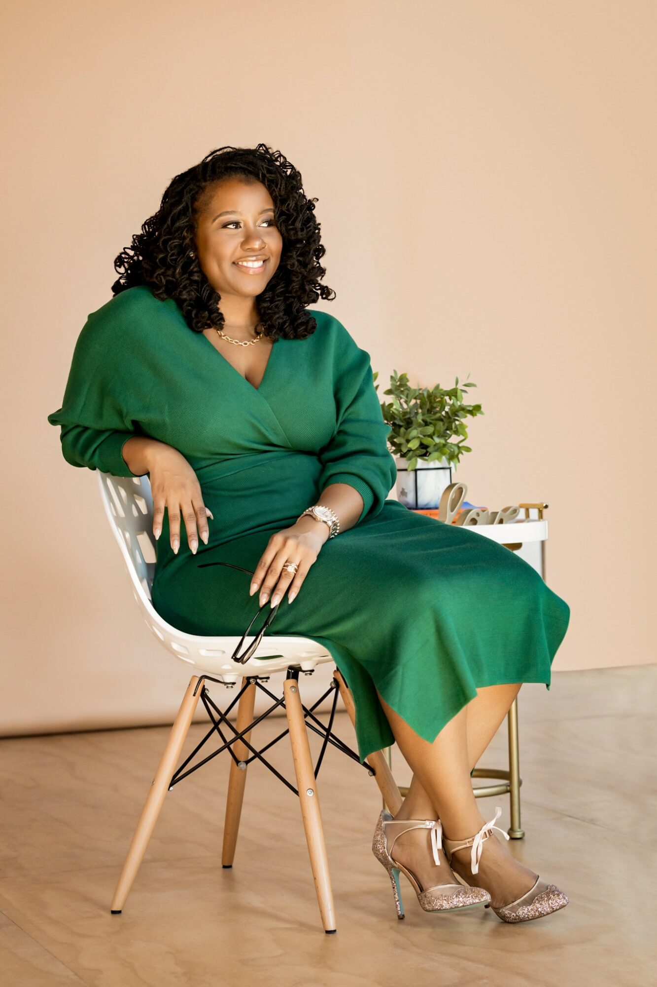 Woman with curly hair in a green dress sitting on a chair, smiling, with a small table and plant behind her.